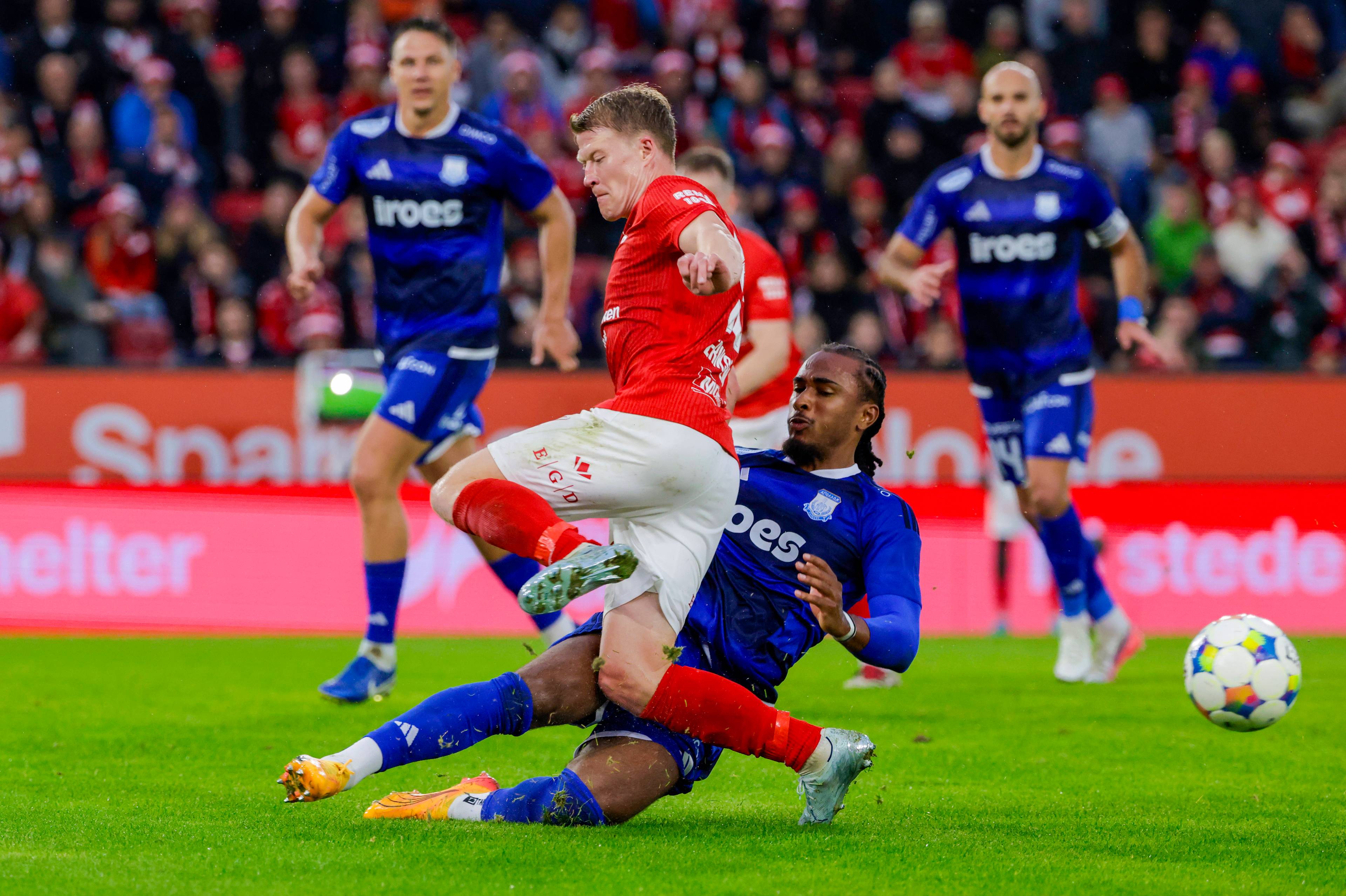 Fotballspiller i rød drakt takles av motspiller i blått under en kamp. Publikum i bakgrunnen. (Bildebeskrivelsen er laget av en KI-tjeneste)