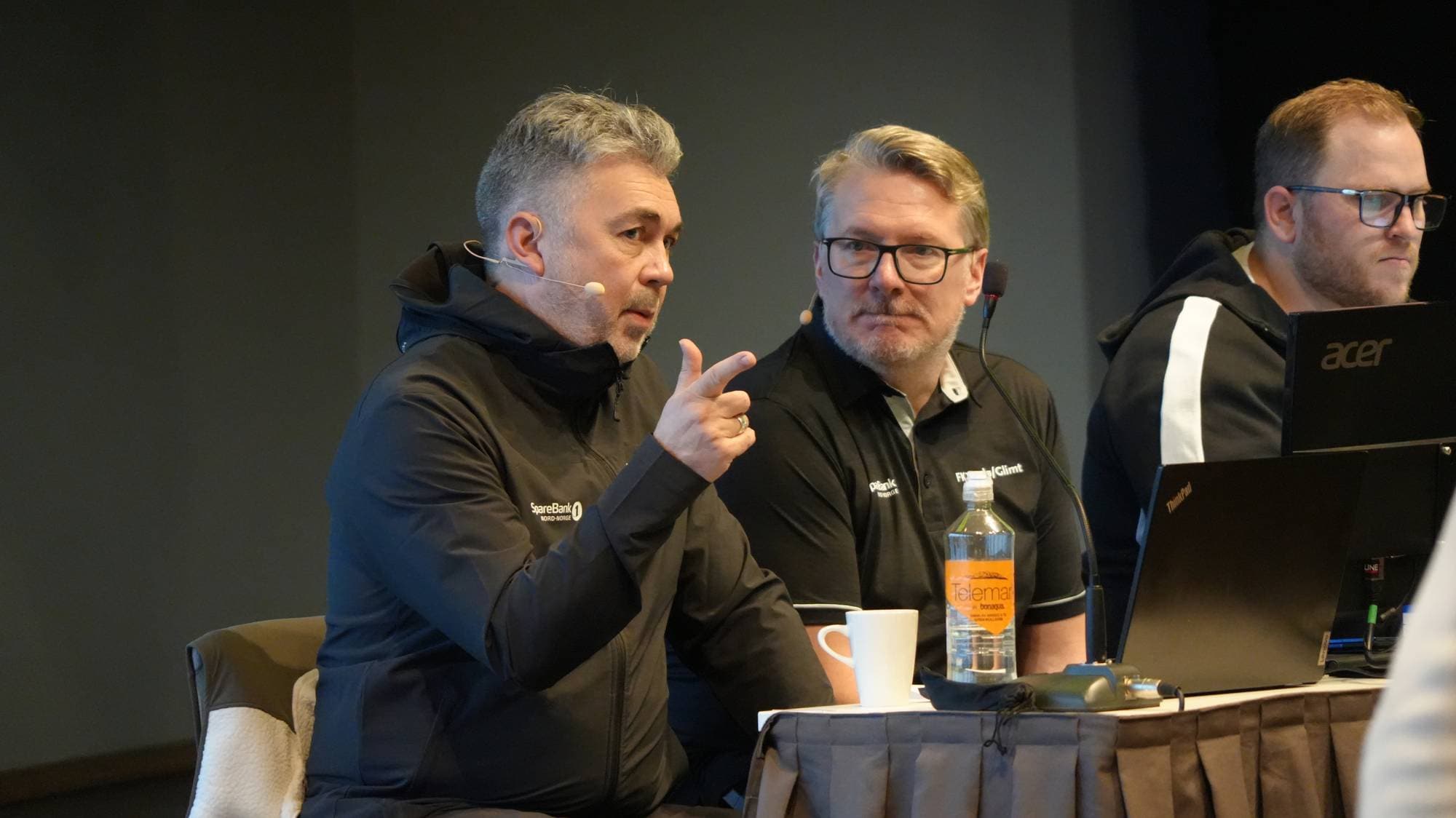 Tre menn sitter ved et bord under et arrangement. En av dem snakker og peker mot publikum. (Bildebeskrivelsen er laget av en KI-tjeneste)