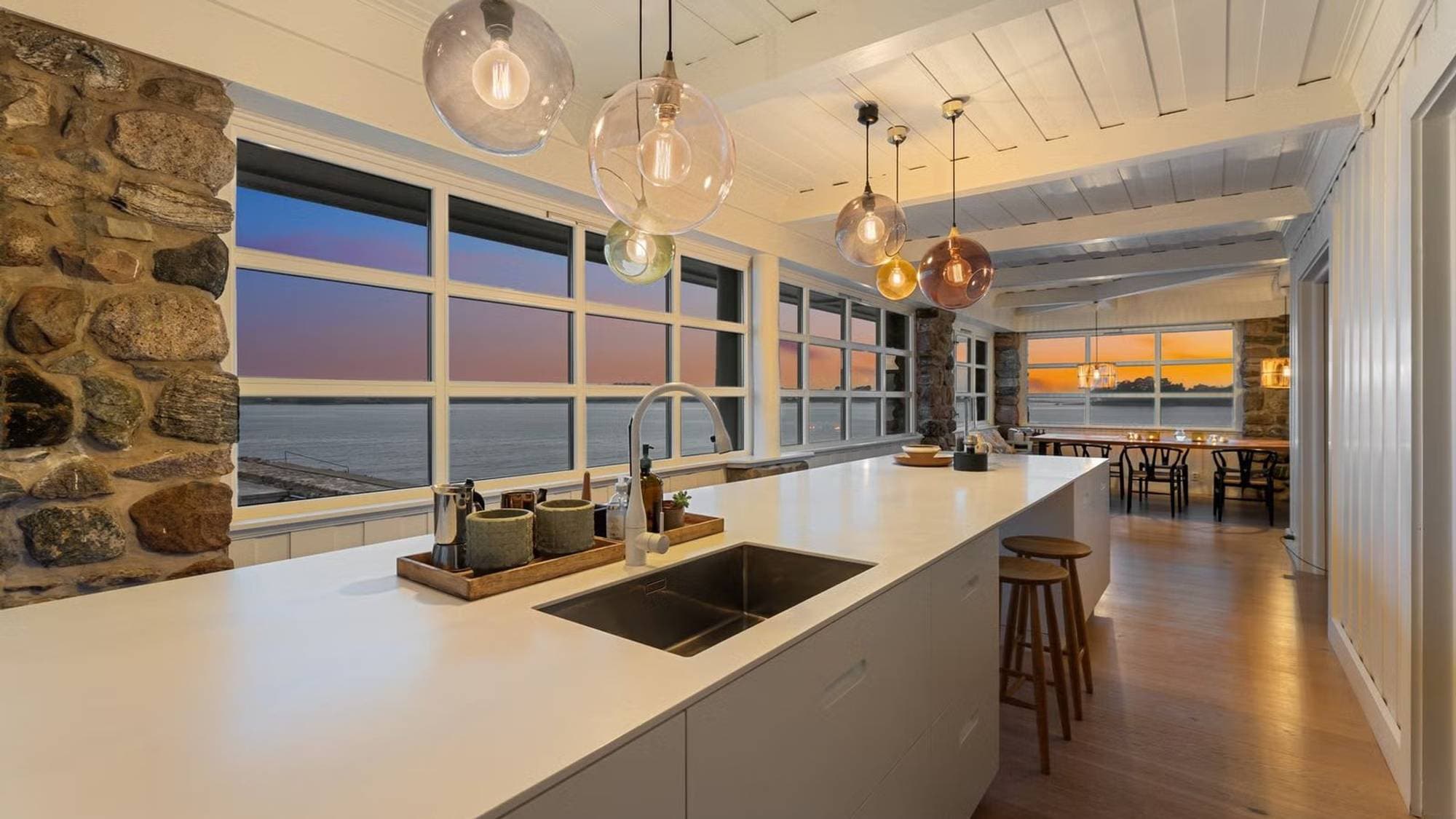 Moderne kjøkken med øy og glasspendler. Store vinduer viser utsikt over sjøen i solnedgang. (Bildebeskrivelsen er laget av en KI-tjeneste)