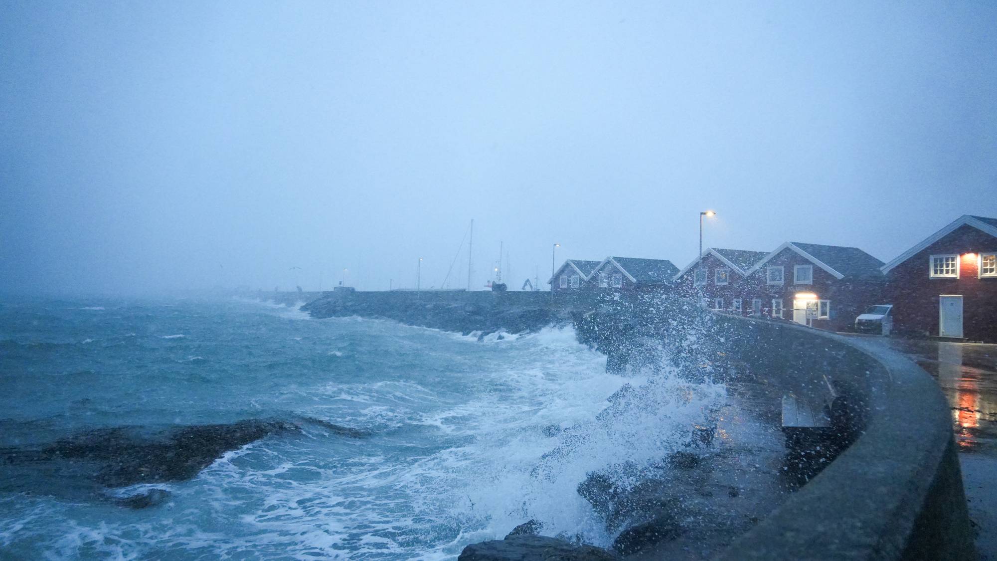Uvær i Bodø i Nordland. Bølgene slår inn mot land over moloen i Bodø og skyller over røde sjøhus. I bakgrunn synes byen i snøvær.