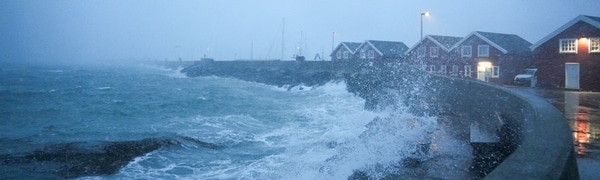 Uvær i Bodø i Nordland. Bølgene slår inn mot land over moloen i Bodø og skyller over røde sjøhus. I bakgrunn synes byen i snøvær.