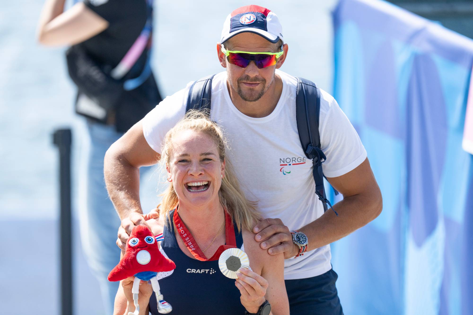 Birgit Skarstein smiler med en medalje og maskot etter Paralympics i Paris. En mann står bak henne. (Bildebeskrivelsen er laget av en KI-tjeneste)