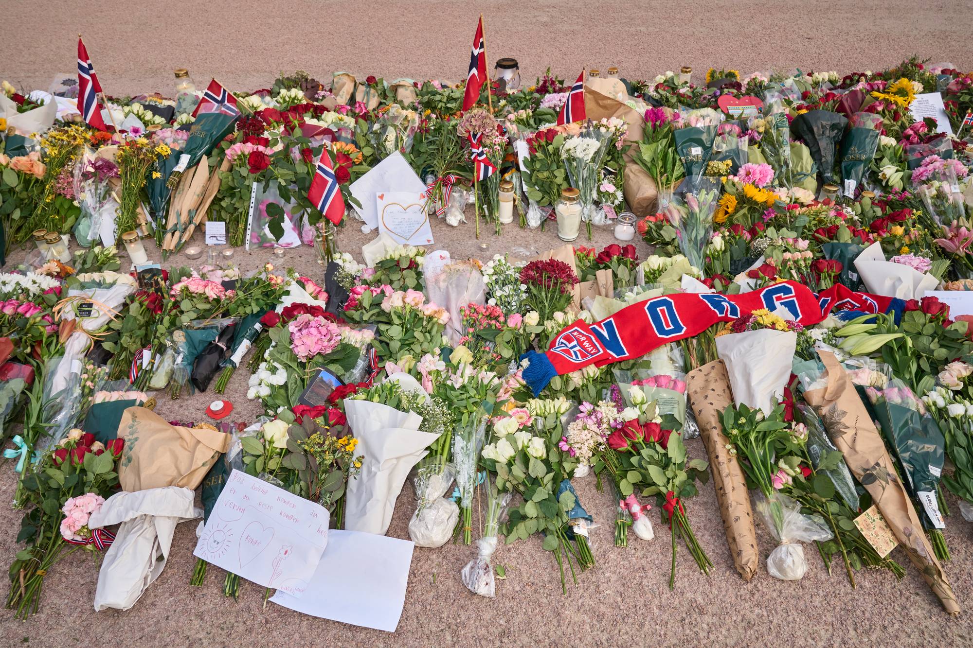 Blomsterhav og norske flagg lagt ned til minne på en åpen plass. Et rødt og hvitt skjerf ligger over. (Bildebeskrivelsen er laget av en KI-tjeneste)