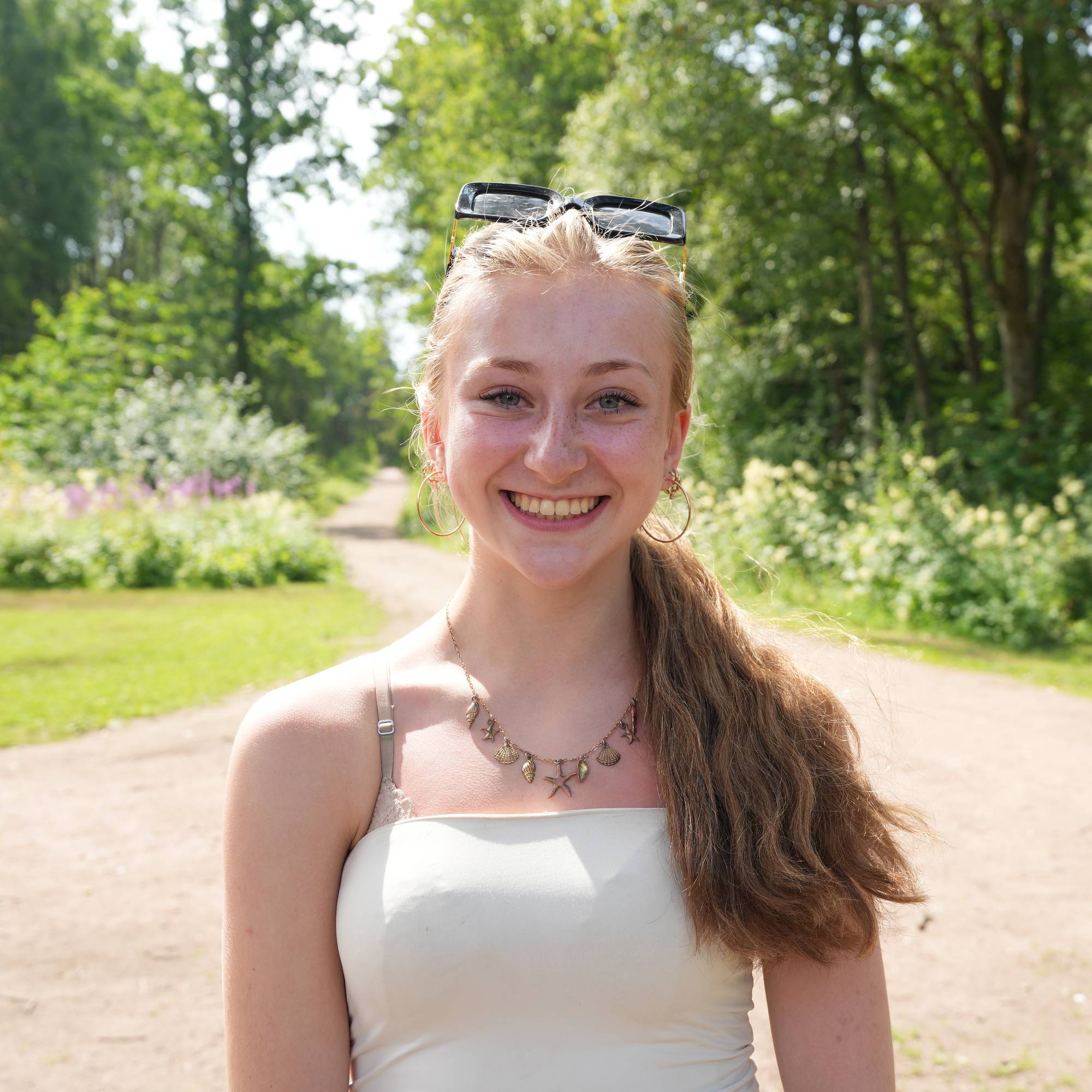 Ung kvinne smiler til kamera på en skogsvei. Hun har solbriller i håret og et smykke med anheng.