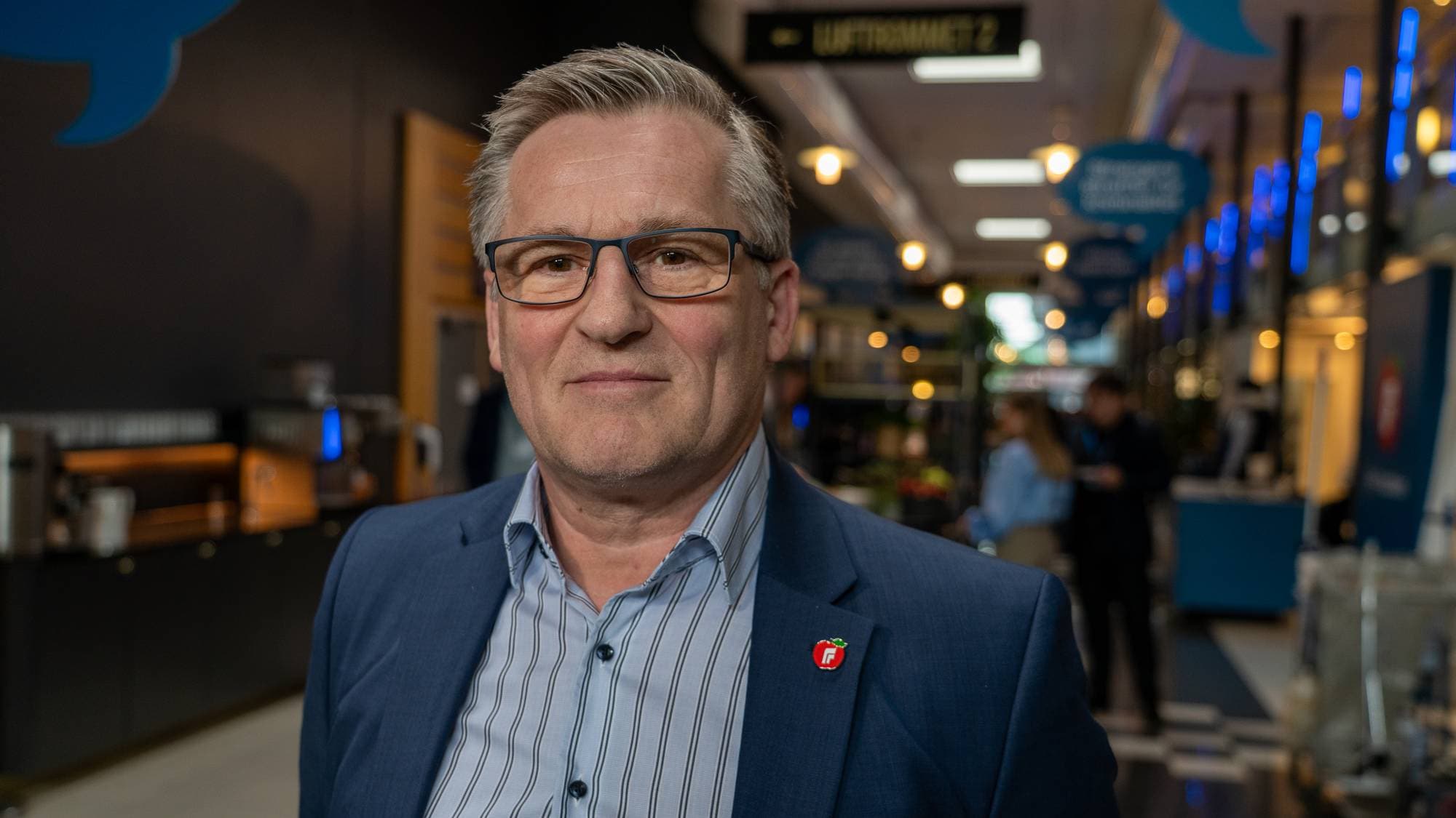 En mann med briller og mørkeblå dress står og ser mot kamera. Han har på seg en stripete skjorte og et rødt merke på reversen. Bak ham er en gang med lys og mennesker i bakgrunnen. (Bildebeskrivelsen er laget av en KI-tjeneste)