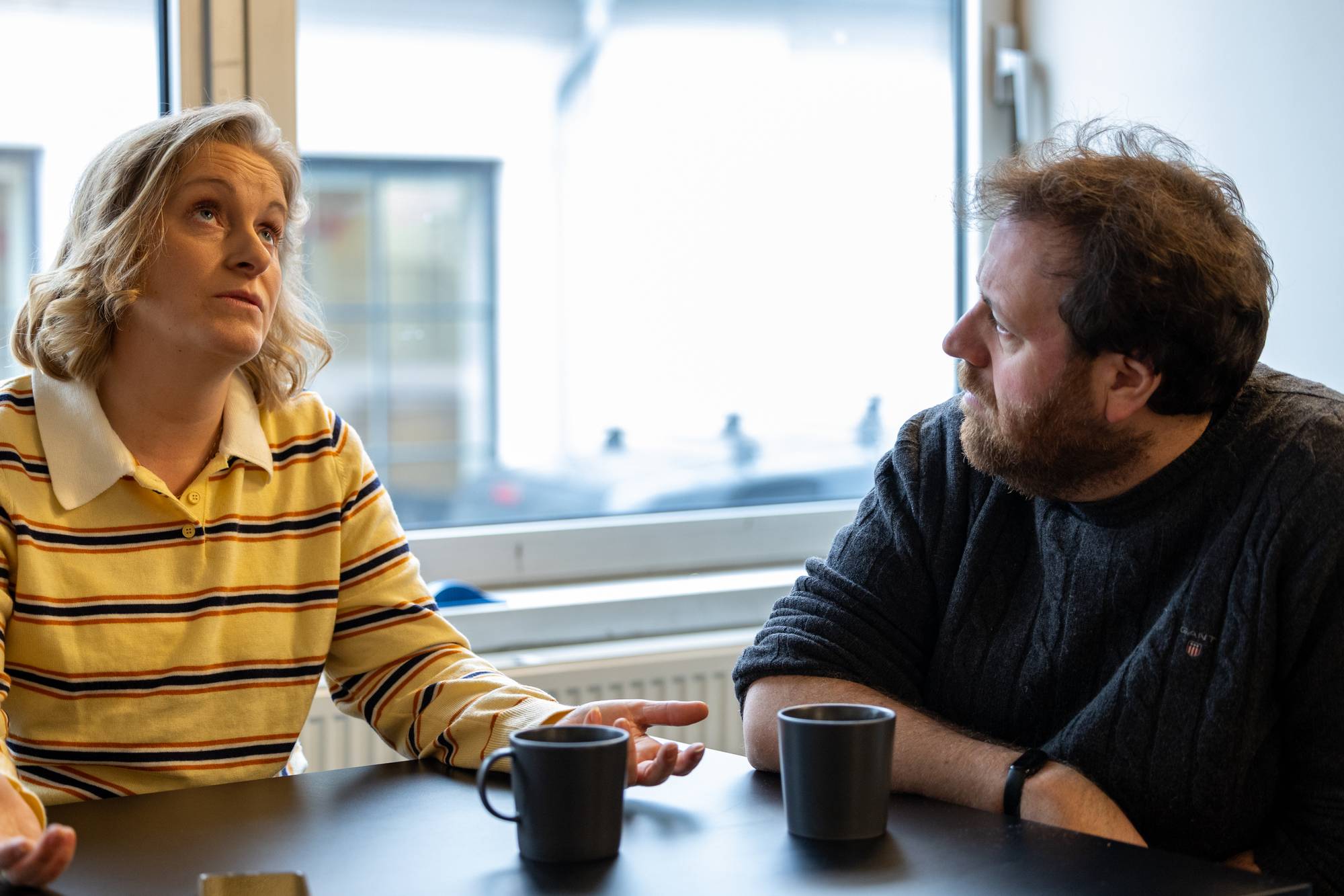 Tuva og Ronny sitter inne foran et vindu. Tuva har lyst hår, og har på seg en gul, stripete genser. Ronny har brunt hår og skjegg, og har på seg en mørk ullgenser. Ronny ser på Tuva som snakker.