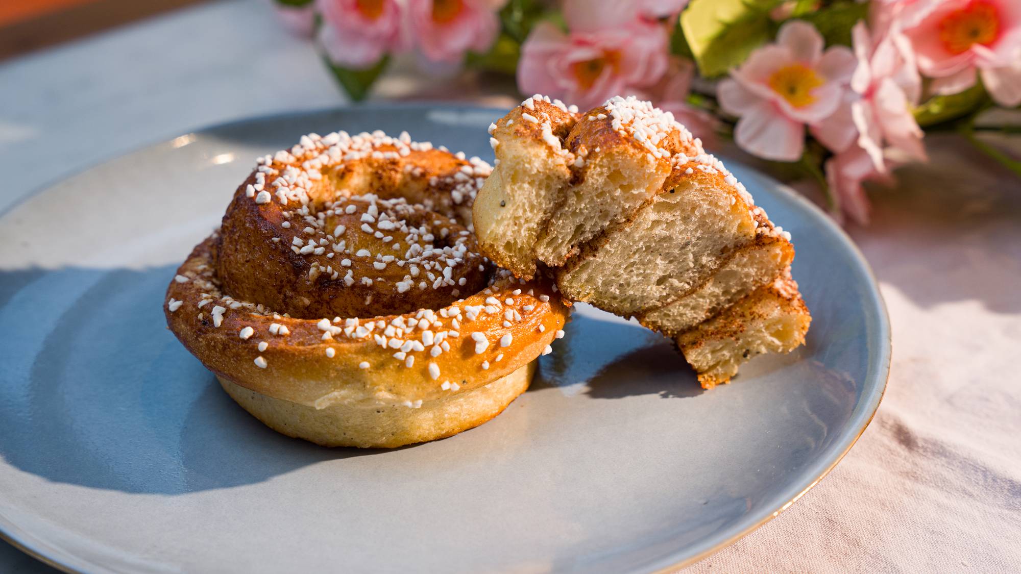 En kanelbolle med sukker strøs på en lys blå tallerken. Den ene delen av kanelbollen er brutt av og viser det indre sukker- og kanelfyllet.