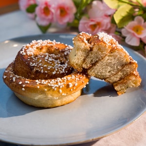 Et kanelbolle med sukker strøs på en lys blå tallerken. Den ene delen av kanelbollen er brutt av og viser det indre sukker- og kanelfyllet.
