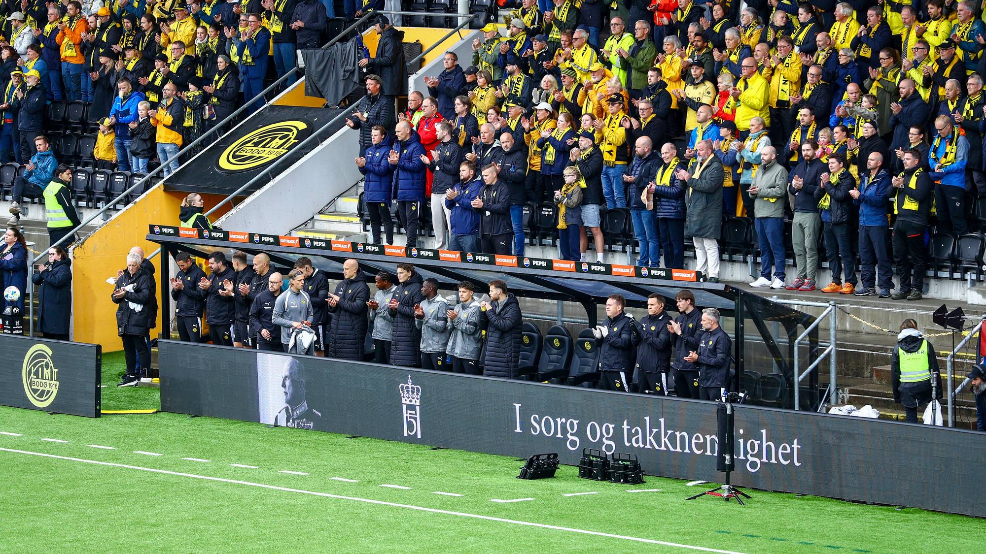 Fotballspillere og publikum står langs banen ved en minnemarkering med teksten I sorg og takknemlighet. (Bildebeskrivelsen er laget av en KI-tjeneste)