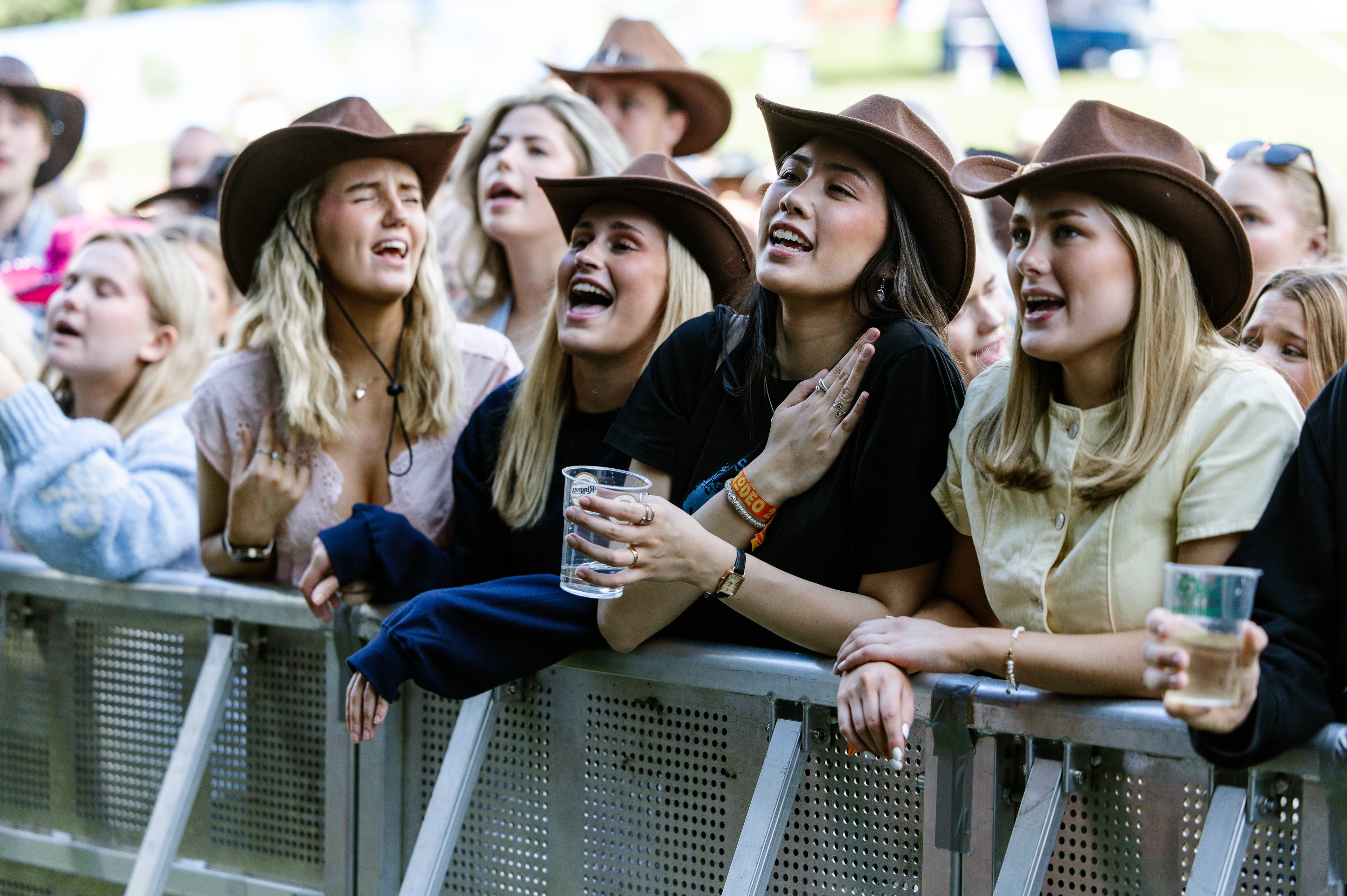 En gruppe unge kvinner med cowboyhatter synger med på musikk under en utendørs konsert. De står tett inntil et sperregitter og holder drikke. Bildet viser en folkemengde som nyter en festival. (Bildebeskrivelsen er laget av en KI-tjeneste)