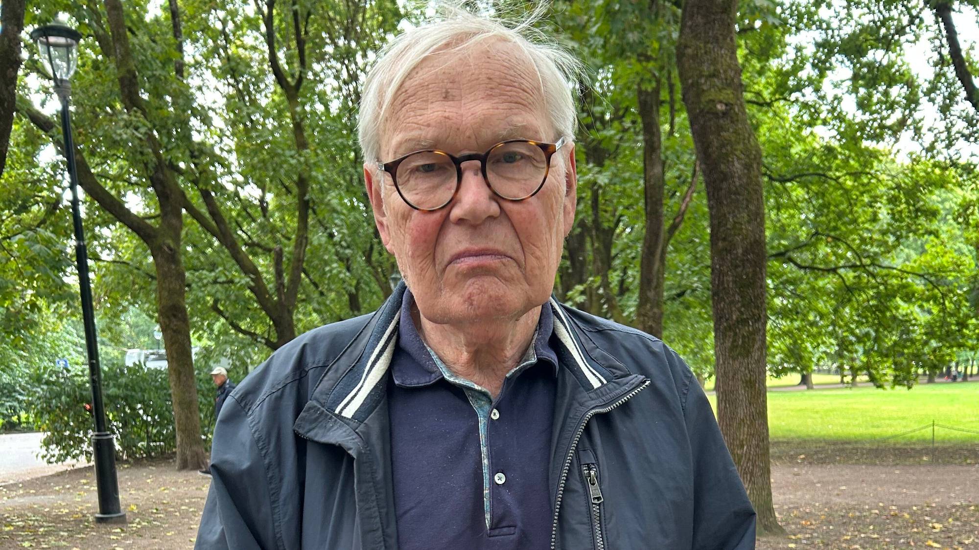 Eldre mann med briller og mørk jakke står i en park. (Bildebeskrivelsen er laget av en KI-tjeneste)