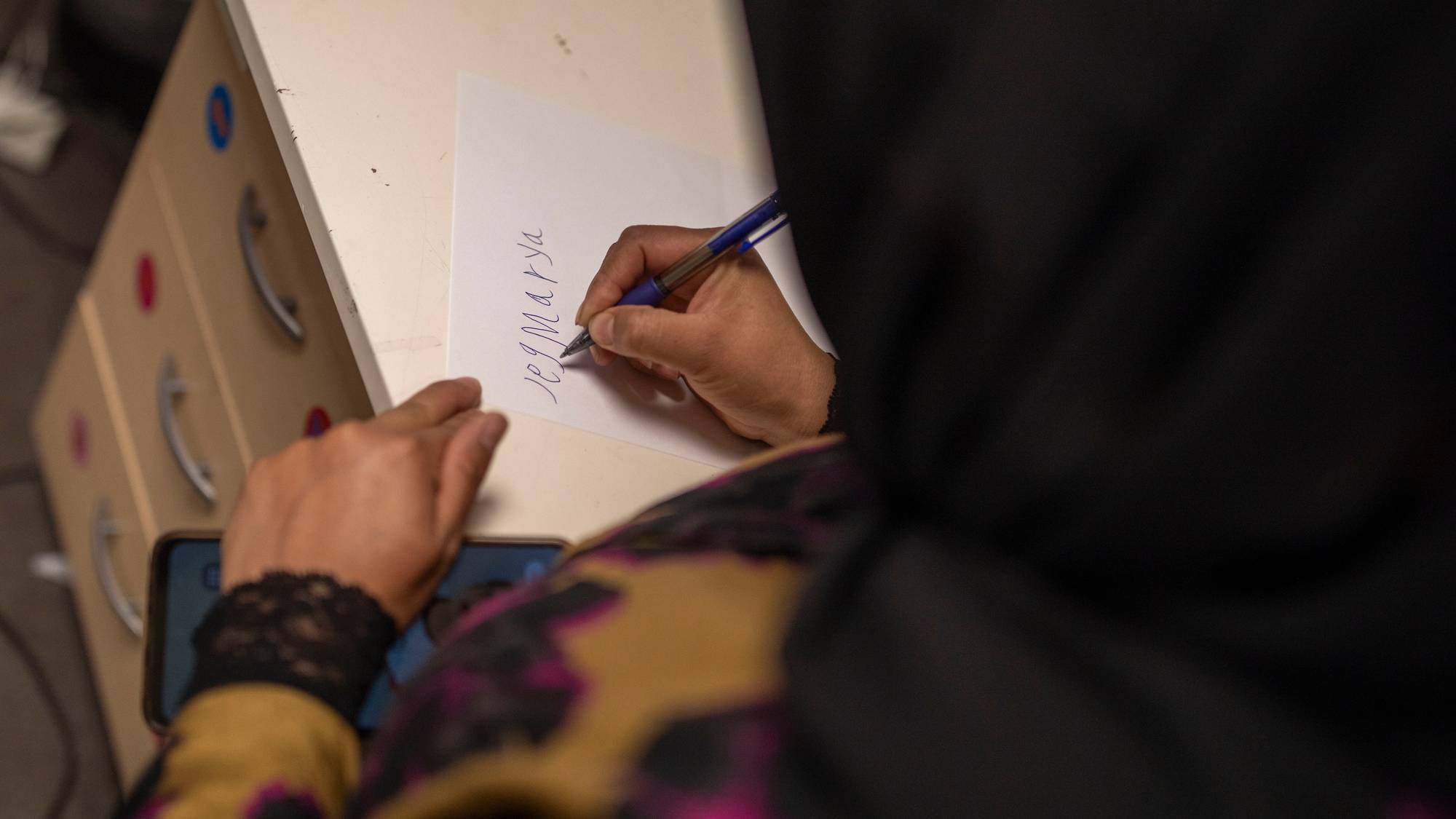 Marya skriver navnet sitt på et ark. Man ser ikke ansiktet hennes. Bildet er tatt bakfra