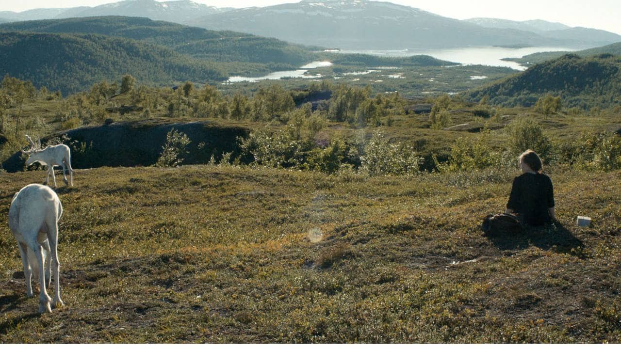 Fra filmen Sameblod. På vidda: Rein til venste, jente sittende til høyre . Vann og fjell der framme