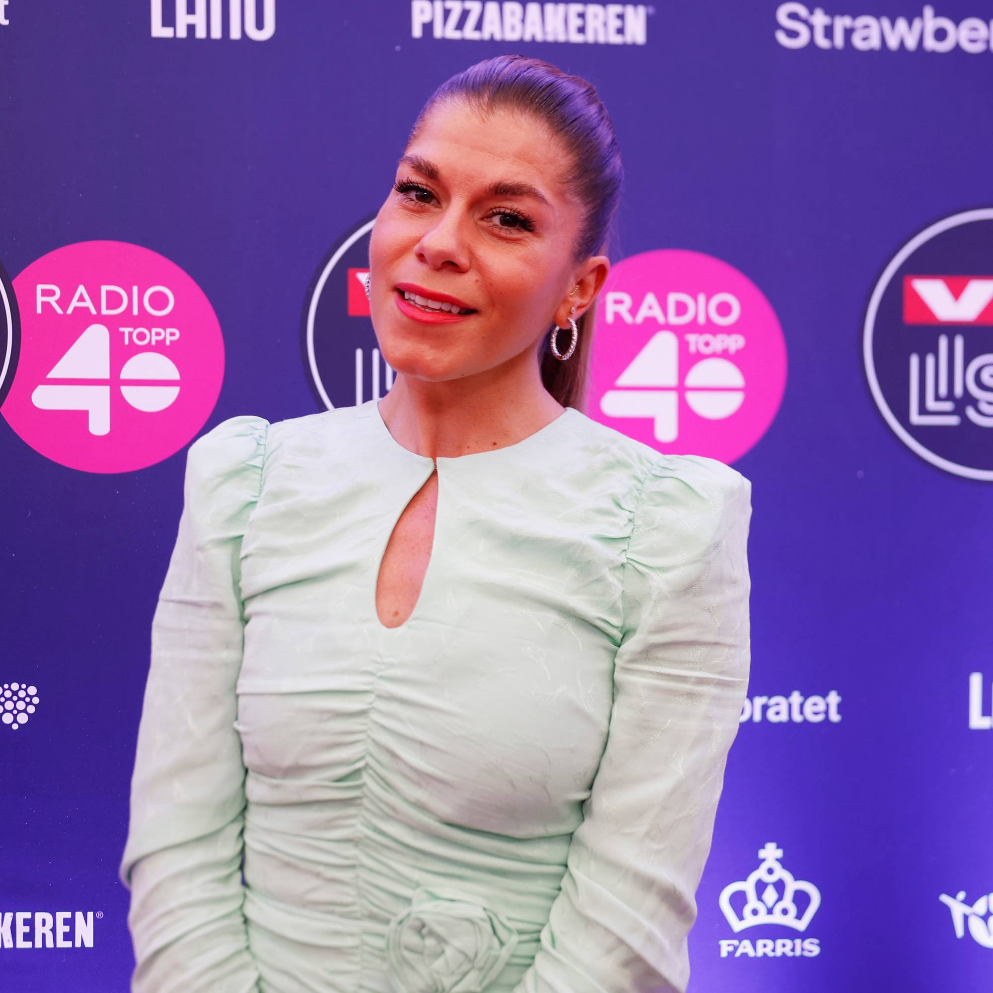 En kvinne med lys mintgrønn kjole og hestehale står foran en lilla bakgrunn. Bak henne vises flere logoer, blant annet VG Lista og Radio Topp 40. Kvinnen smiler mot kameraet. (Bildebeskrivelsen er laget av en KI-tjeneste)