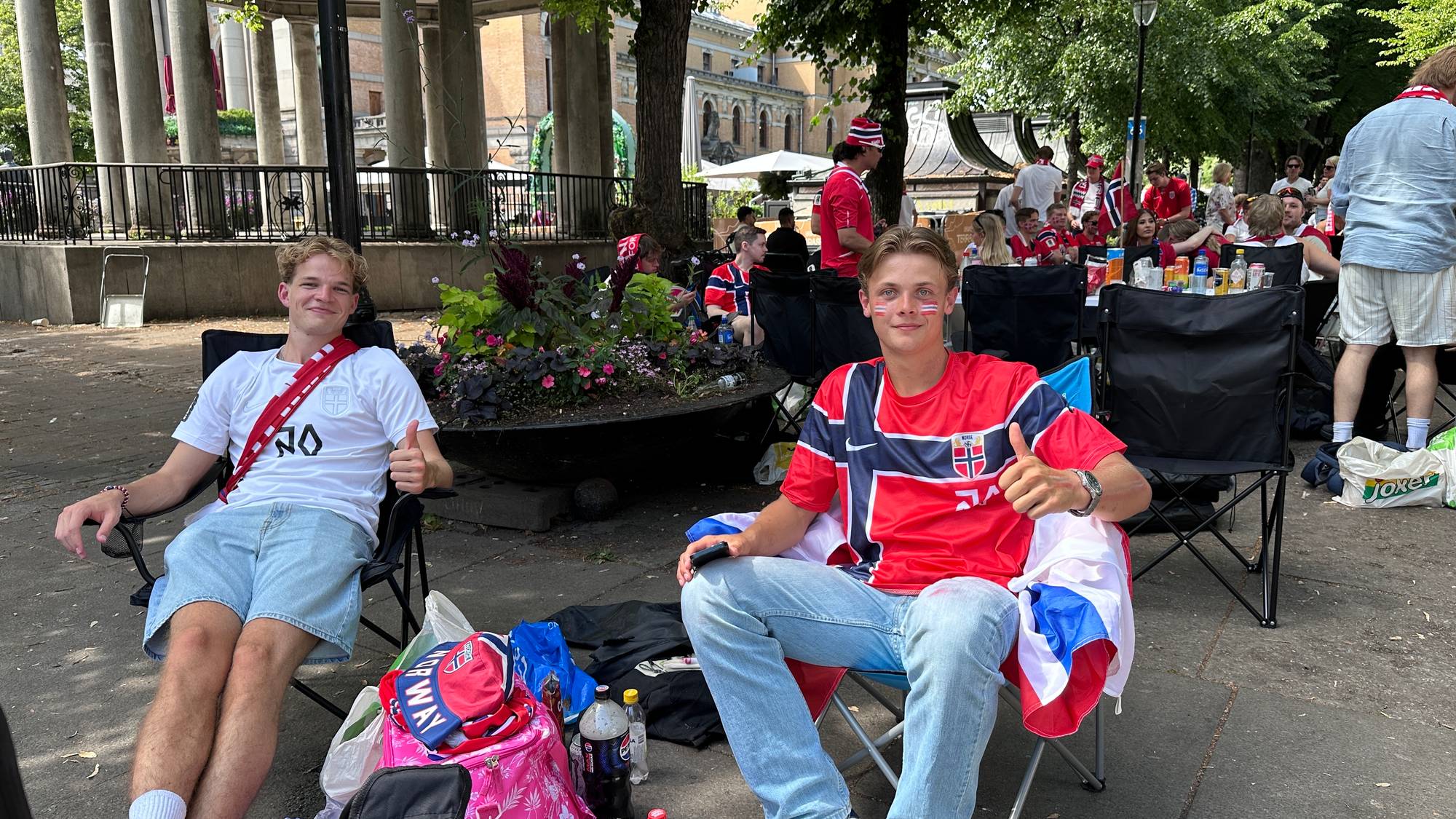 To unge menn i norske landslagsklær sitter i campingstoler i en park og gir tommel opp. (Bildebeskrivelsen er laget av en KI-tjeneste)