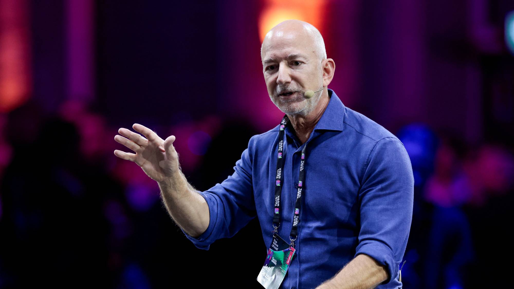 Jeff Bezos snakker på en scene under en konferanse, kledd i blå skjorte med adgangskort rundt halsen. (Bildebeskrivelsen er laget av en KI-tjeneste)