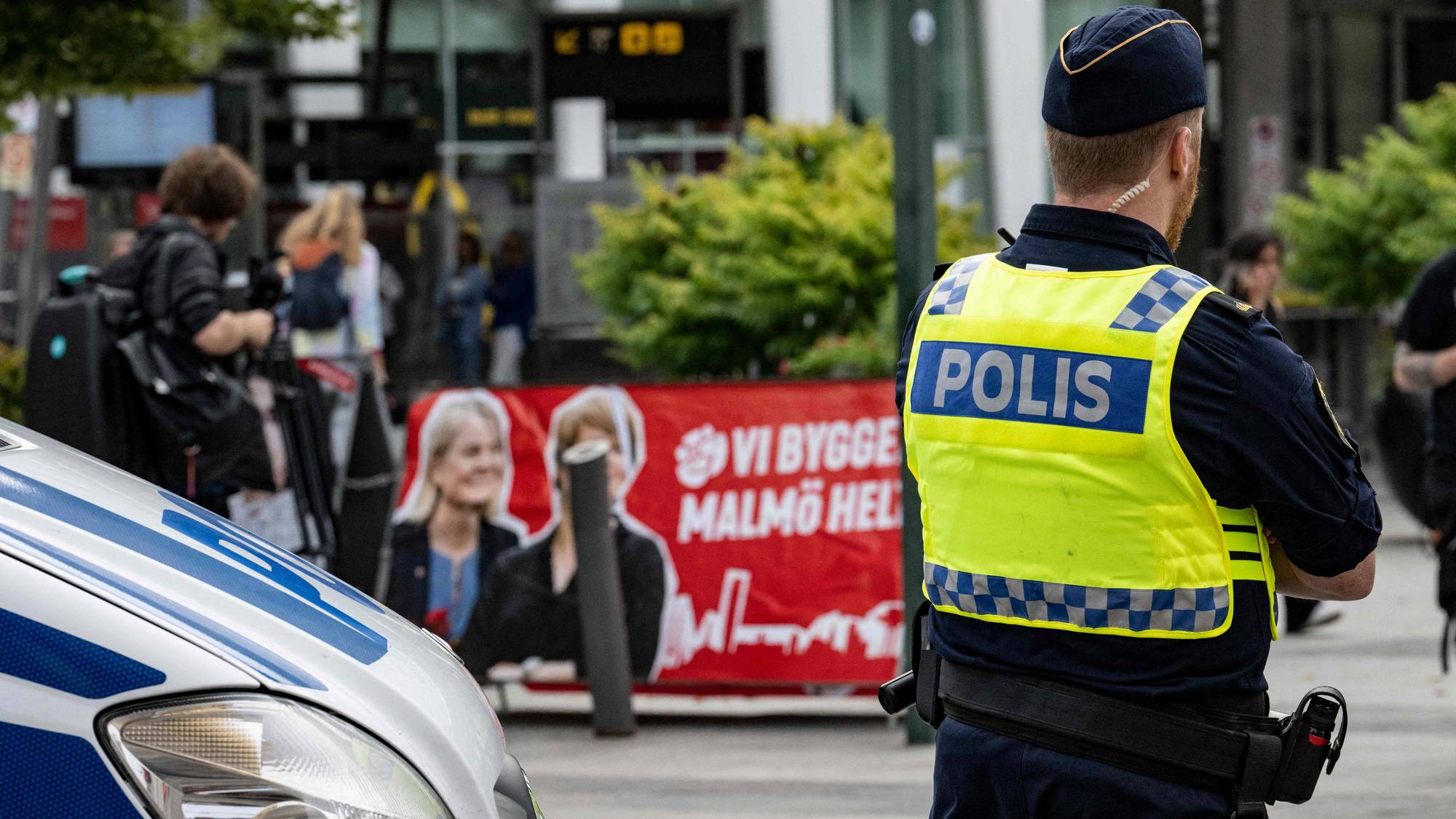 Svensk politibetjent står ved en politibil med en valgplakat i bakgrunnen. (Bildebeskrivelsen er laget av en KI-tjeneste)