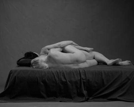 En naken mann med langt hvitt hår ligger sidelengs i senga bak en naken kvinne med mørkt hår og hode på en pute