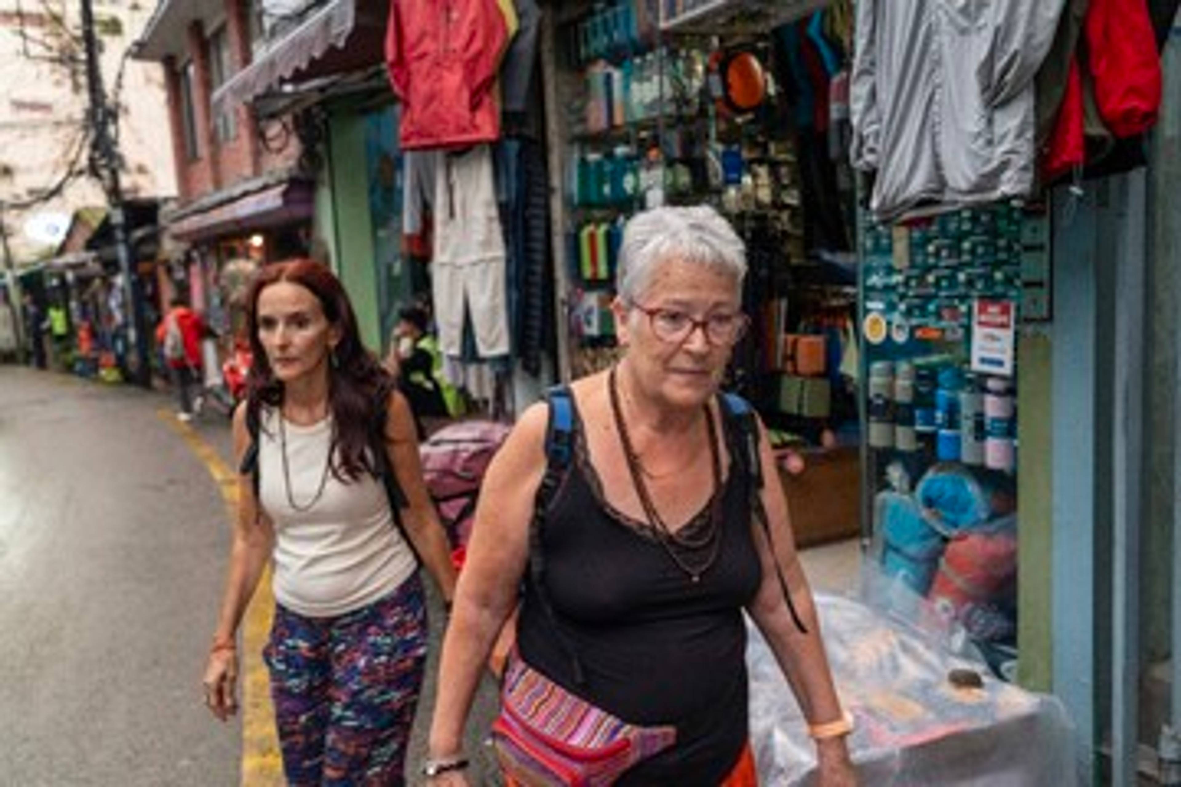 Turistar i Katmandu – Gråhåret dame går i ein gate med butikkar. Klede henger på fasadane til butikkane.