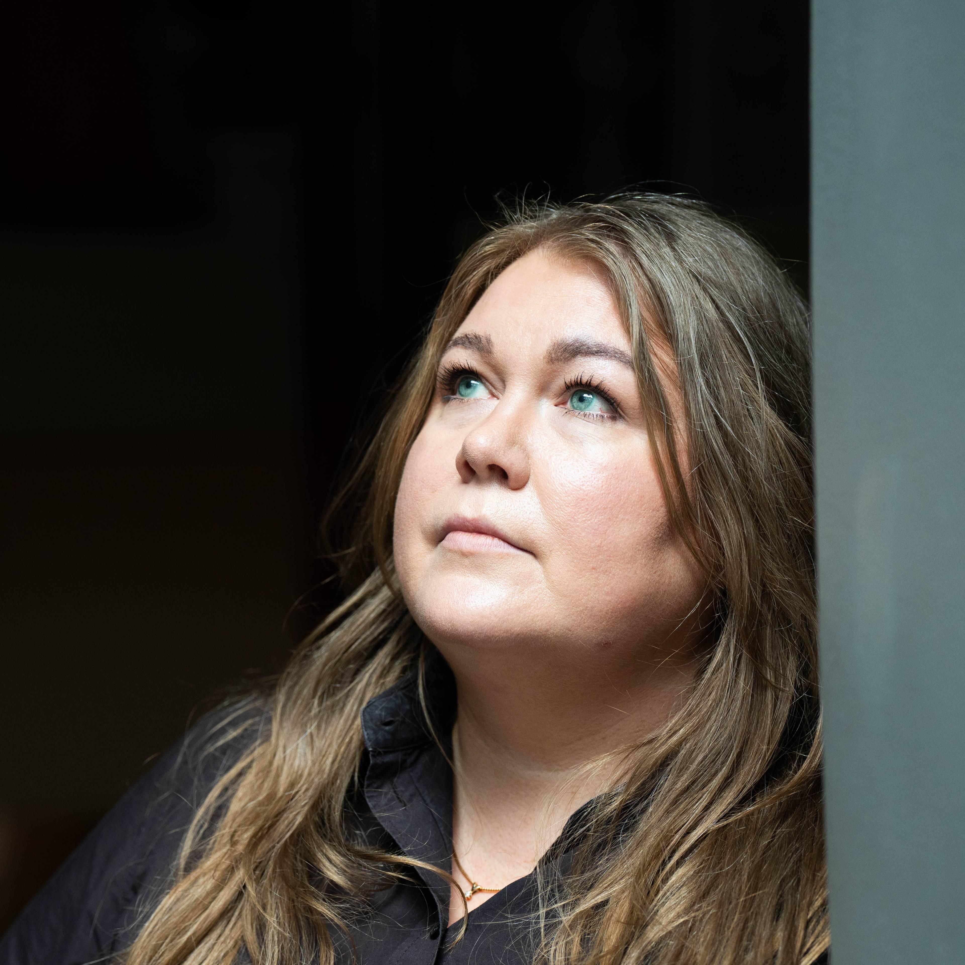 En kvinne med brunt hår, svart genser og store blå øyne ser opp mot venstre.