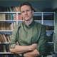 Frode Fjerdingstad / NRK En person står med armene kryssede foran en bokhylle fylt med bøker. Personen har på seg en grønn skjorte. Omgivelsene inkluderer planter og en stabel papir. Belysningen i rommet er naturlig og gir et varmt preg.