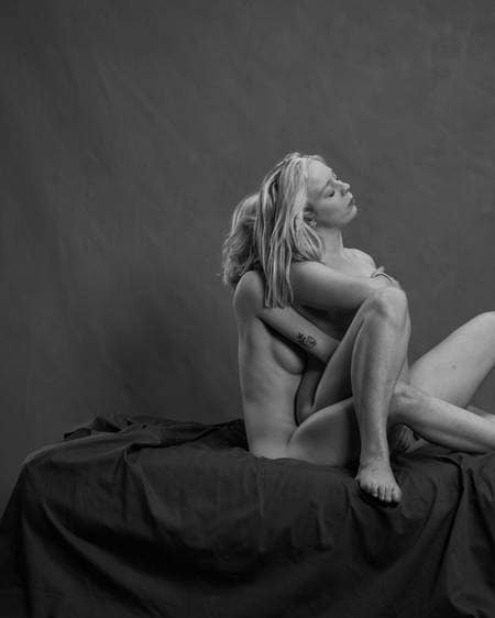 To nakne damer med lyst hår sitter bak hverandre i senga. De holder rundt hverandre og lukker øynene