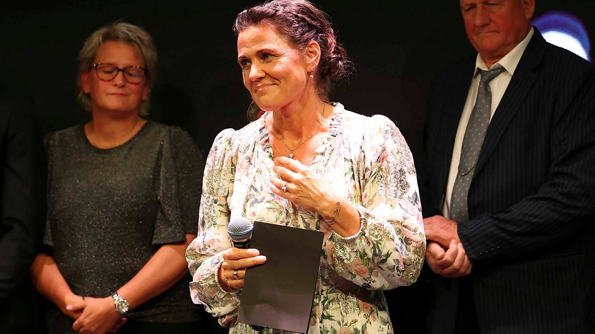 Kvinne med blomstrete kjole står med mikrofon og papirer. En mann og en kvinne står ved siden av. (Bildebeskrivelsen er laget av en KI-tjeneste)