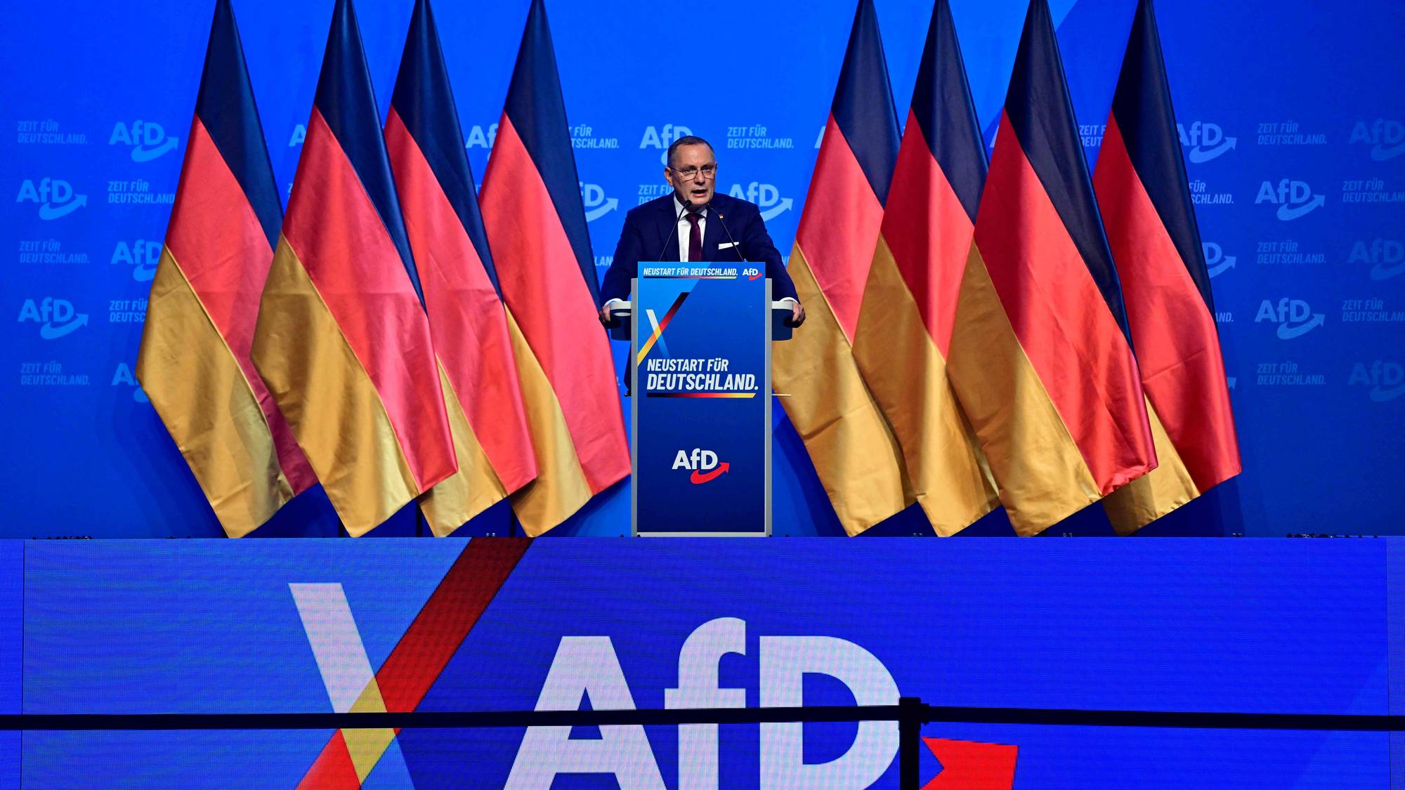 Tino Chrupalla taler fra en talerstol foran tyske flagg og AfD-logoer under et partimøte. (Bildebeskrivelsen er laget av en KI-tjeneste)