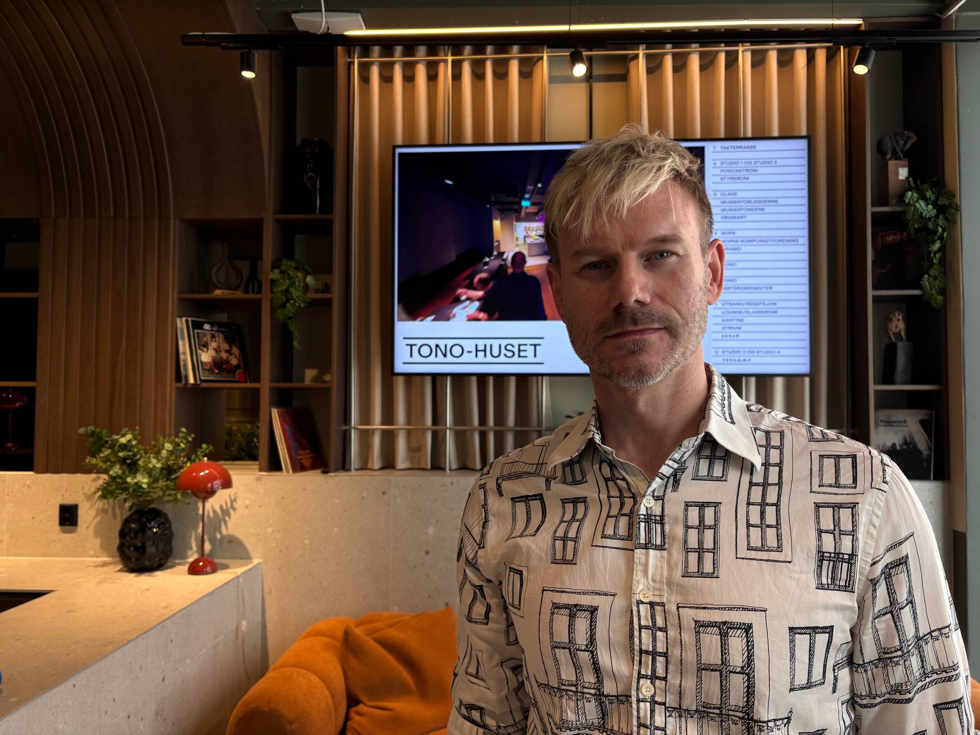 Mann med kvit skjorte med vindauge ser inn i kamera. Sofa og TV i bakgrunnen