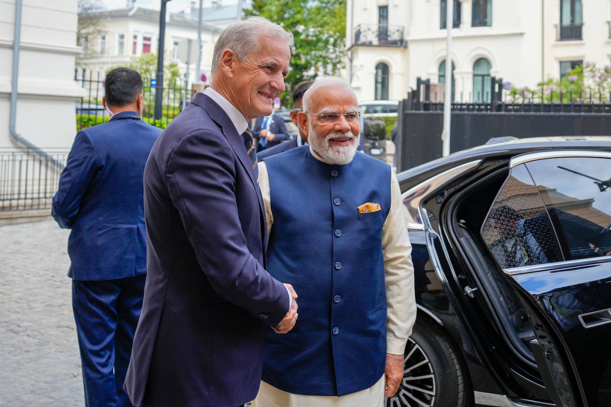 Statsminister Jonas Gahr Støre hilser på Indias statsminister Narendra Modi. Modi er iført en blå vest over lys skjorte og kurta. De står foran en svart bil med døren åpen.