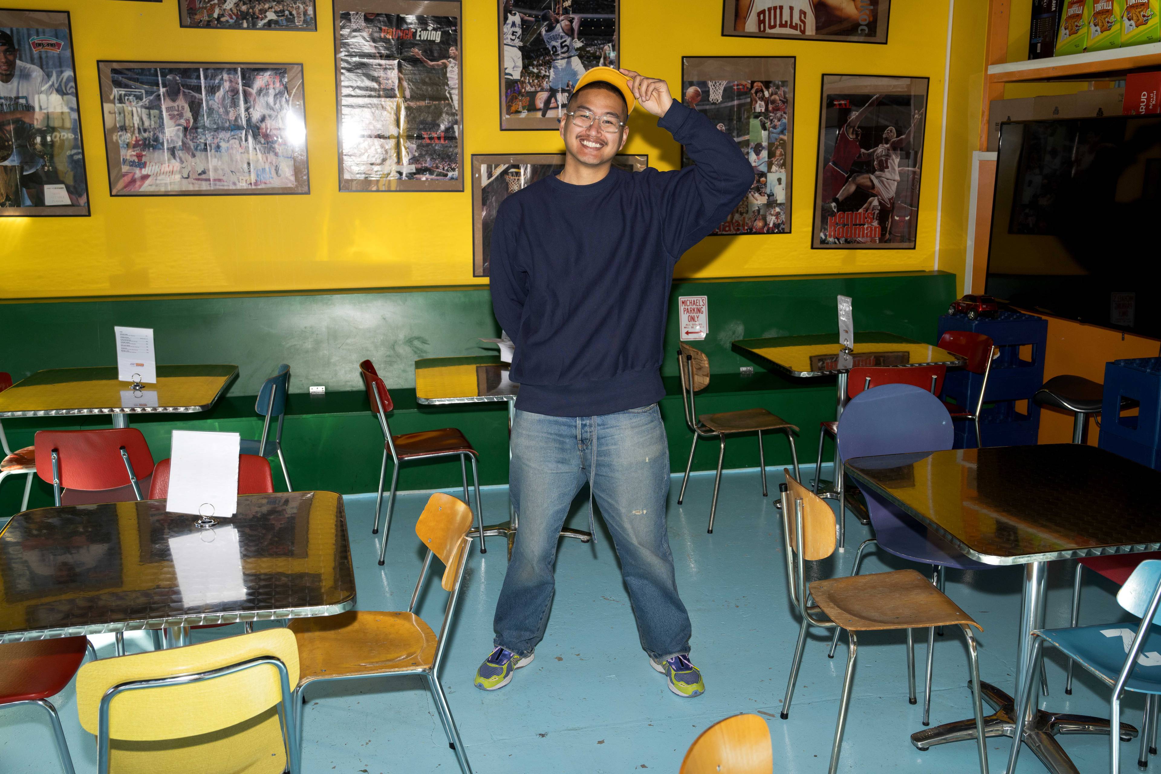 Mike står midt i rommet på Mike's Corner, løfter på capsen og smiler til kamera. Rundt han står bord og stoler, og på veggen henger det ulike basketballplakater