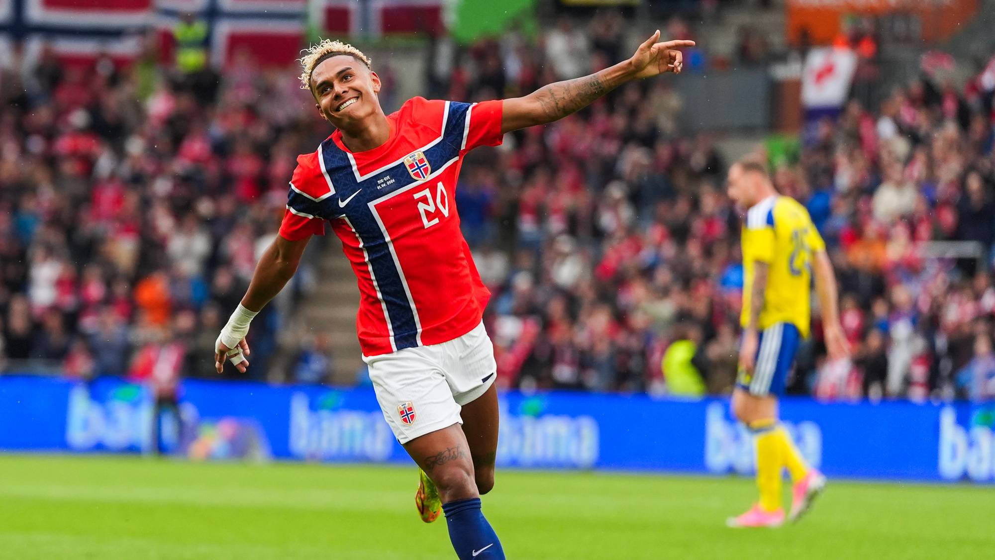 En fotballspiller i rød og blå drakt feirer et mål på en gressbane. I bakgrunnen er det publikum og en annen spiller i gul drakt. (Bildebeskrivelsen er laget av en KI-tjeneste)
