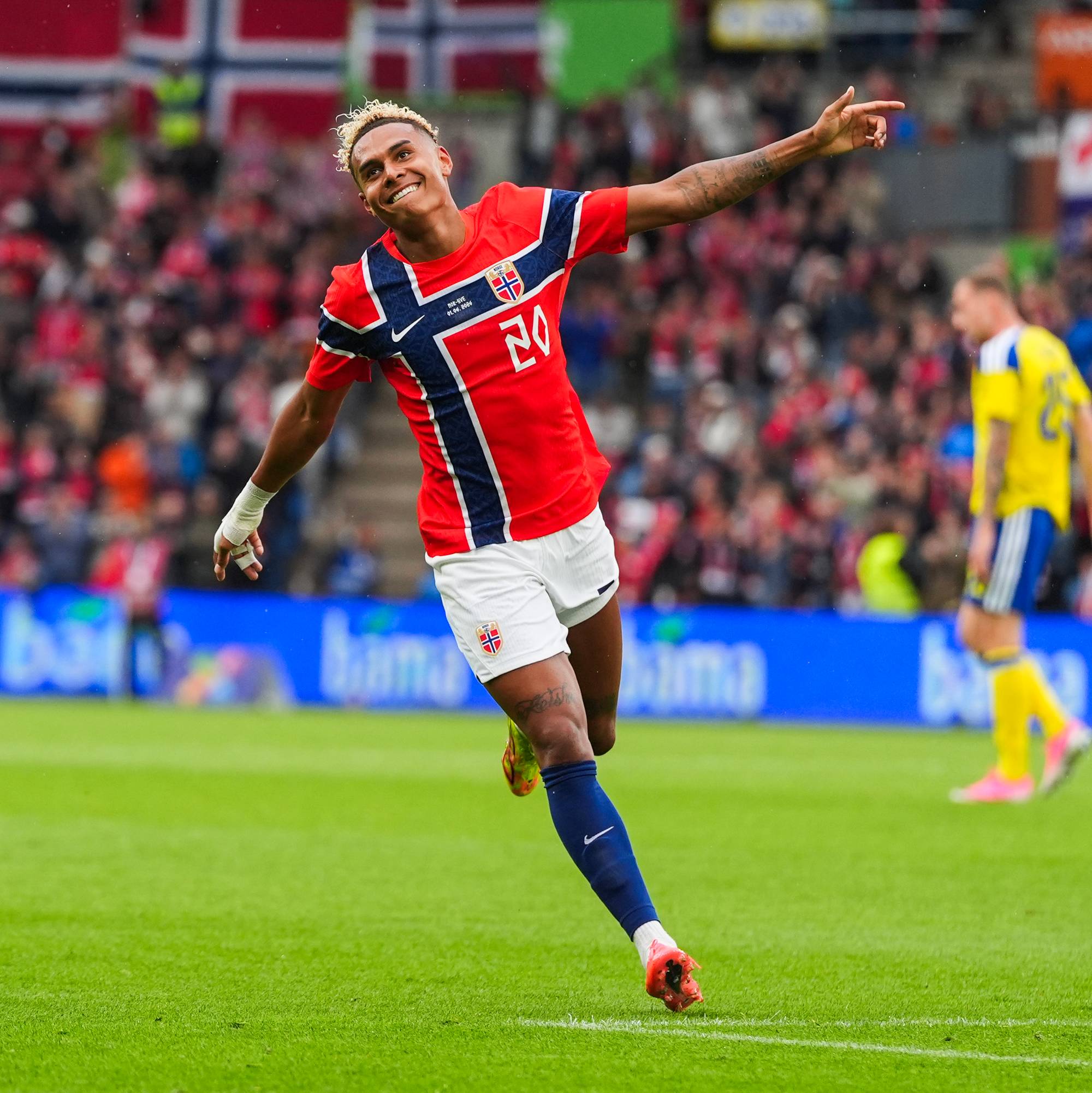 En fotballspiller i rød og blå drakt feirer et mål på en gressbane. I bakgrunnen er det publikum og en annen spiller i gul drakt. (Bildebeskrivelsen er laget av en KI-tjeneste)