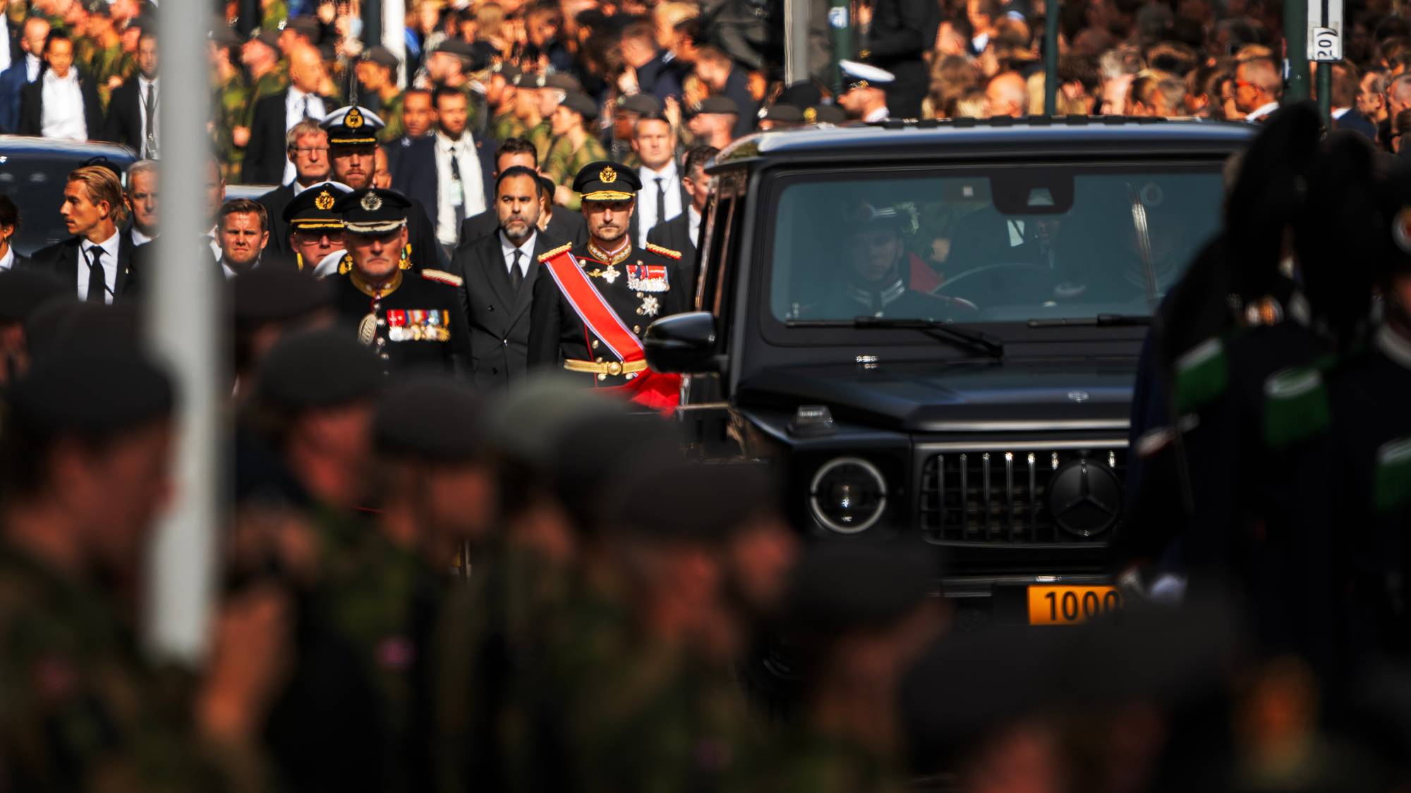 Kronprins Haakon går i militær prosesjon forbi en svart bil i en folkemengde. (Bildebeskrivelsen er laget av en KI-tjeneste)