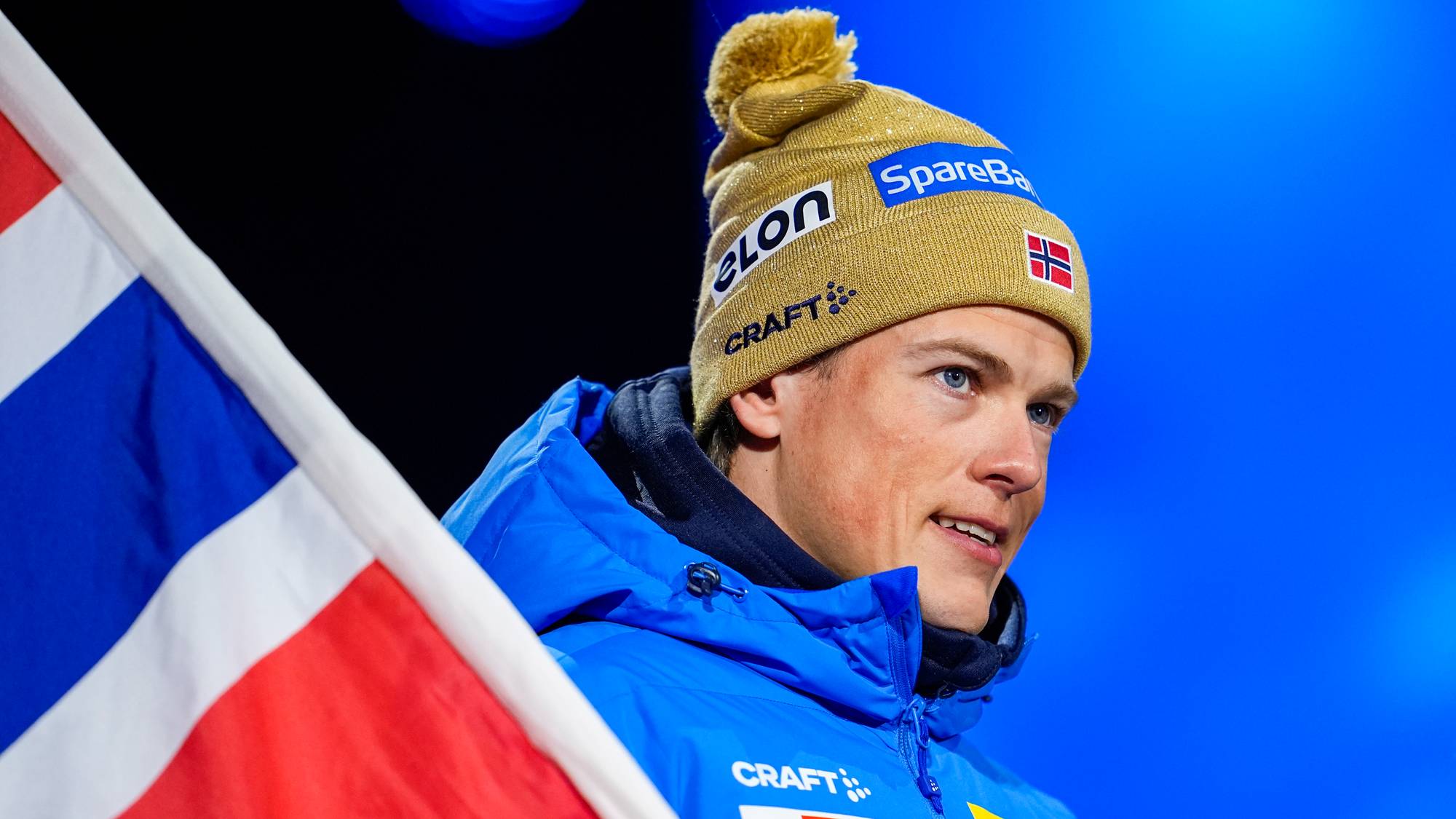 Johannes Thingnes Bø med norsk flagg og gullfarget lue foran blå bakgrunn. (Bildebeskrivelsen er laget av en KI-tjeneste)