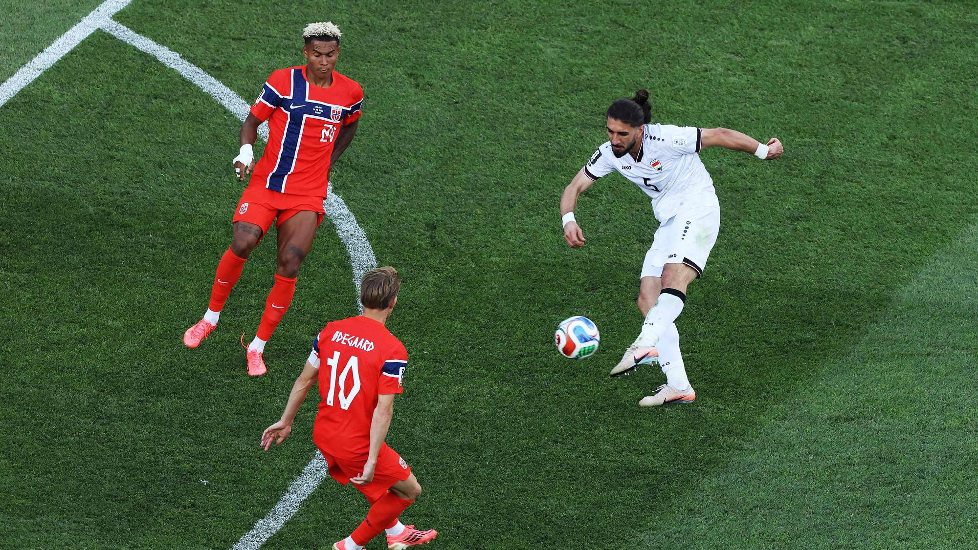 En fotballspiller i hvit drakt sparker ballen. To spillere i røde drakter ser på. De er på en gressbane med hvite linjer. (Bildebeskrivelsen er laget av en KI-tjeneste)