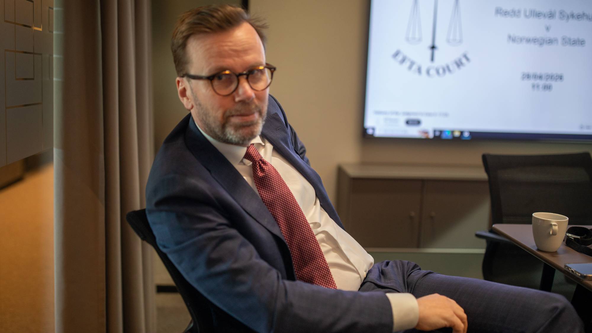 En mann i dress og slips sitter i en stol. Bak ham er en skjerm med et rettsvesen-emblem. Mannen har briller og ser mot kamera. Han sitter i et møterom med et vindu på venstre side. (Bildebeskrivelsen er laget av en KI-tjeneste)