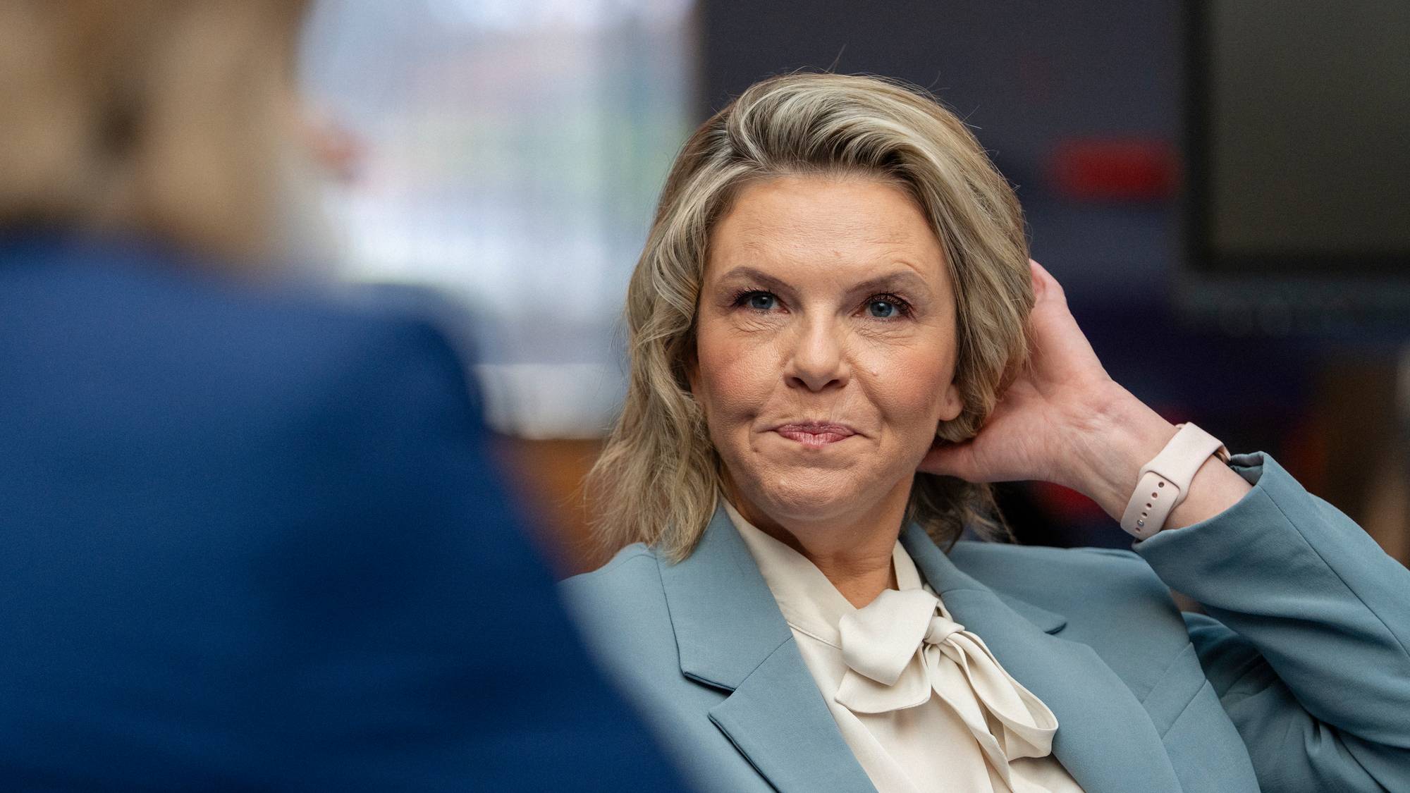 En kvinne med blondt hår, iført en lyseblå dressjakke, ser til høyre. Hun har høyre hånd på bakhodet og en lys klokke på venstre håndledd. En person i blå dress sitter foran henne. (Bildebeskrivelsen er laget av en KI-tjeneste)
