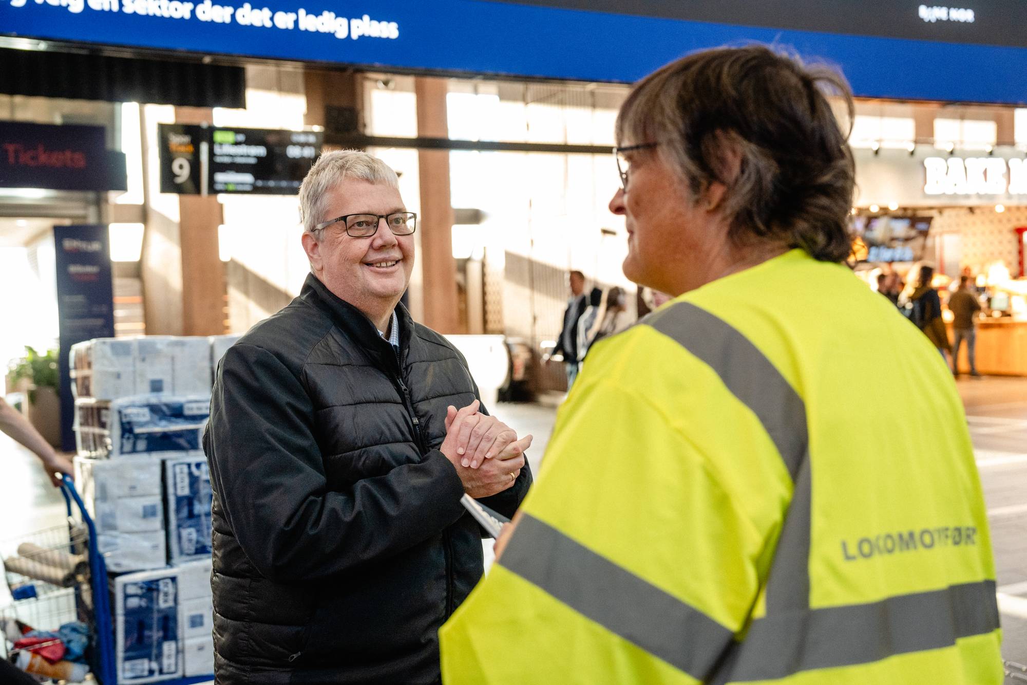 To personer snakker sammen på en togstasjon. En lokomotivfører i gul refleksvest står med ryggen til. (Bildebeskrivelsen er laget av en KI-tjeneste)