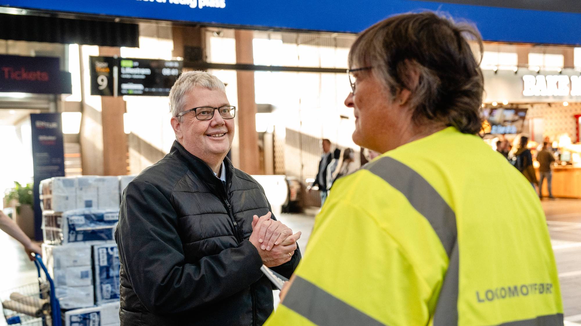 To personer snakker sammen på en togstasjon. En lokomotivfører i gul refleksvest står med ryggen til. (Bildebeskrivelsen er laget av en KI-tjeneste)
