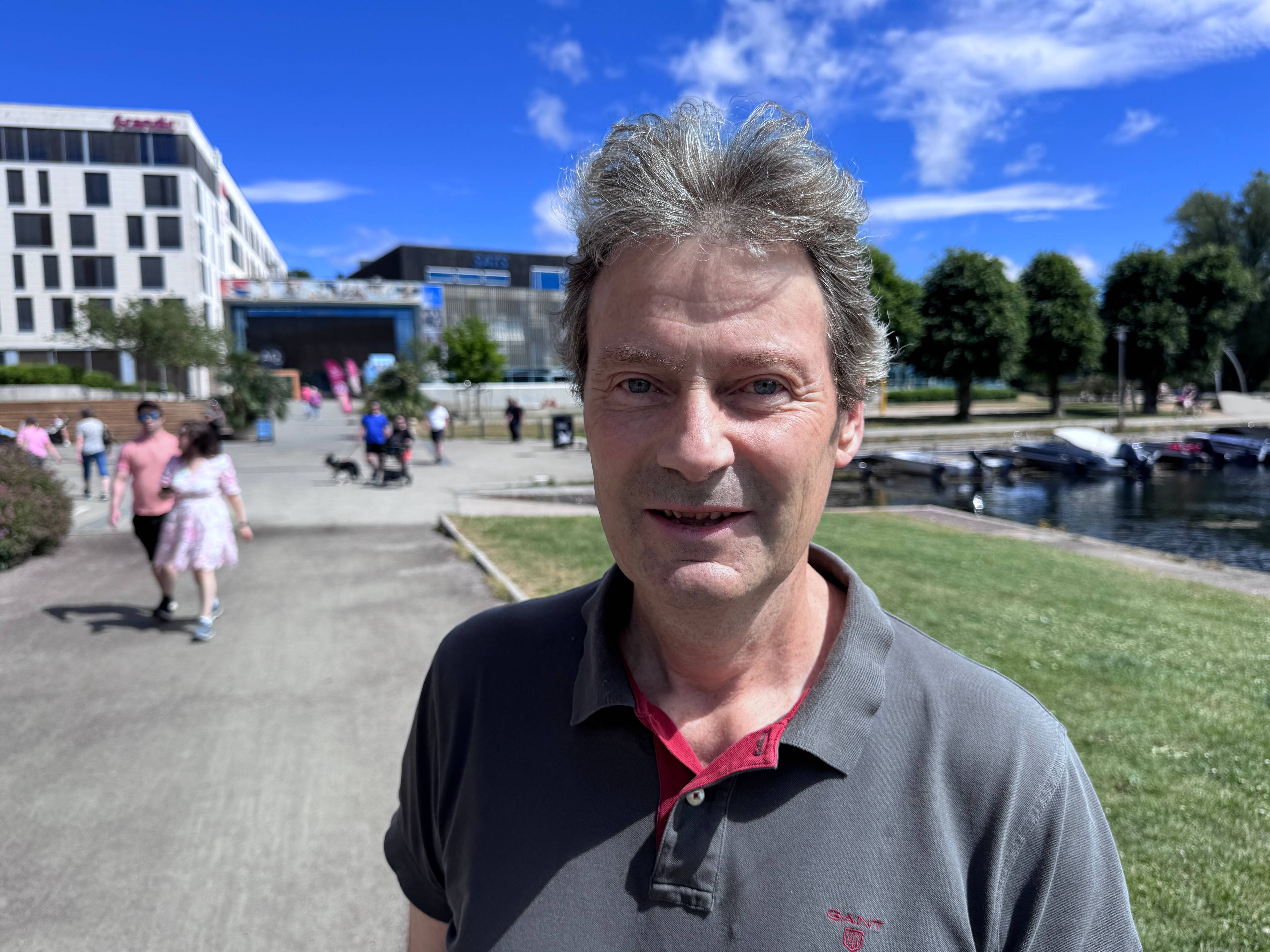 En mann med grå t-skjorte står på en grusvei. I bakgrunnen ser man flere folk og en hvit bygning.