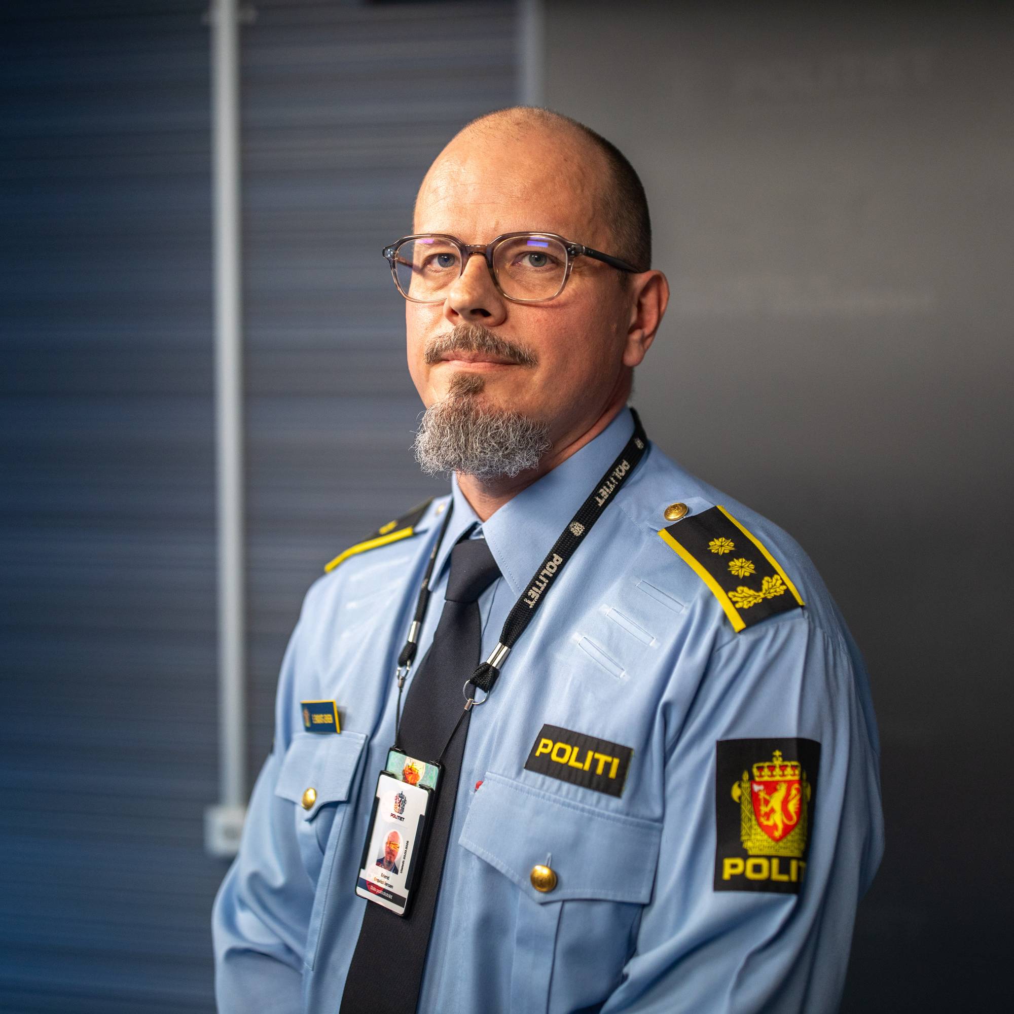 En mann med briller og skjegg ser inn i kamera. Han har på seg en politiuniform.