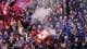 NTB En stor folkemengde med tilskuere ser på en fotballkamp. Flere personer bruker fakler som avgir mye røyk, og et stort supporterflagg er synlig. Tilskuerne har på seg skjerf og klær i lagets farger, og noen har ansiktsmaling. (Bildebeskrivelsen er laget av en KI-tjeneste)