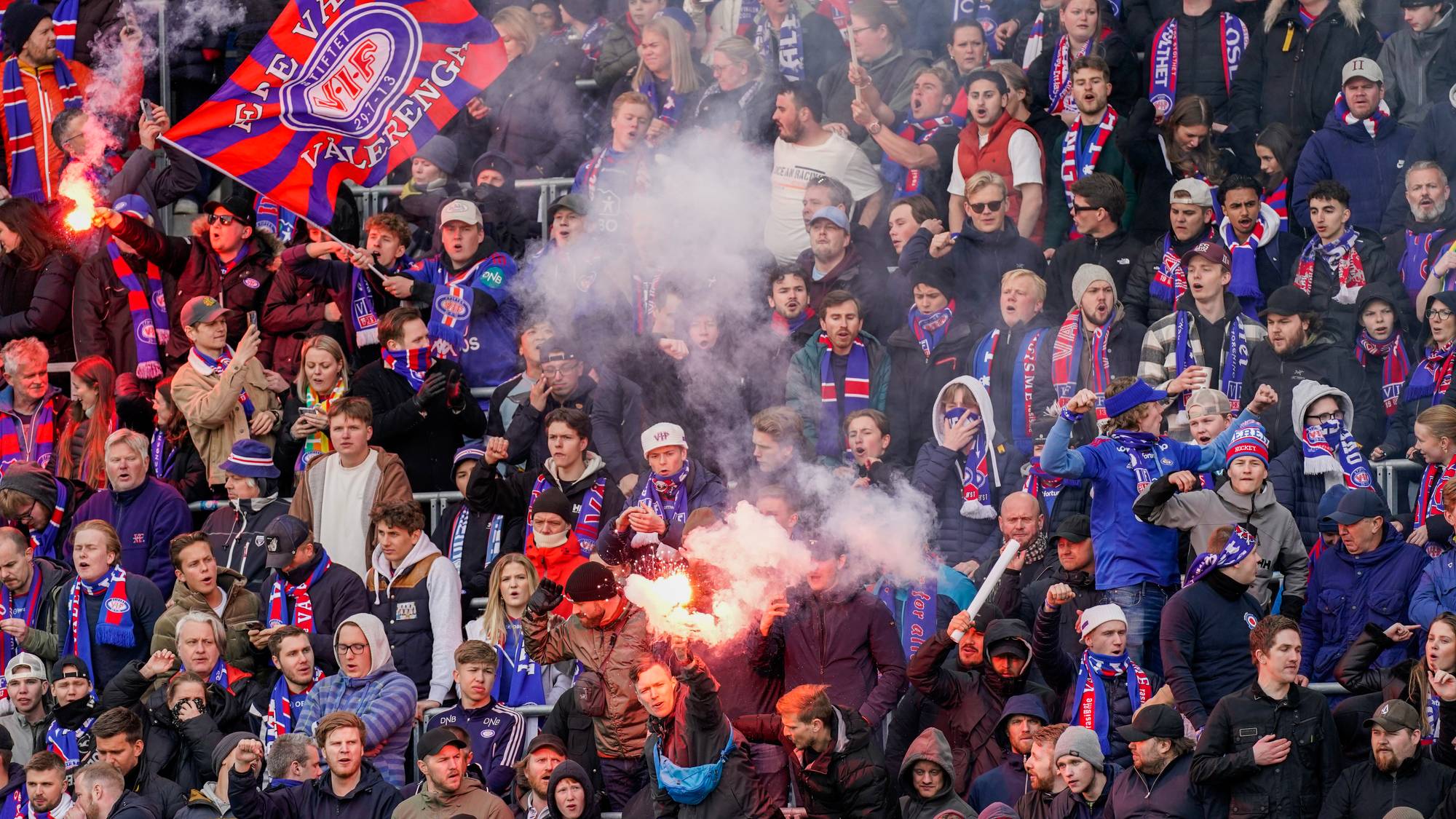 En stor folkemengde med tilskuere ser på en fotballkamp. Flere personer bruker fakler som avgir mye røyk, og et stort supporterflagg er synlig. Tilskuerne har på seg skjerf og klær i lagets farger, og noen har ansiktsmaling. (Bildebeskrivelsen er laget av en KI-tjeneste)