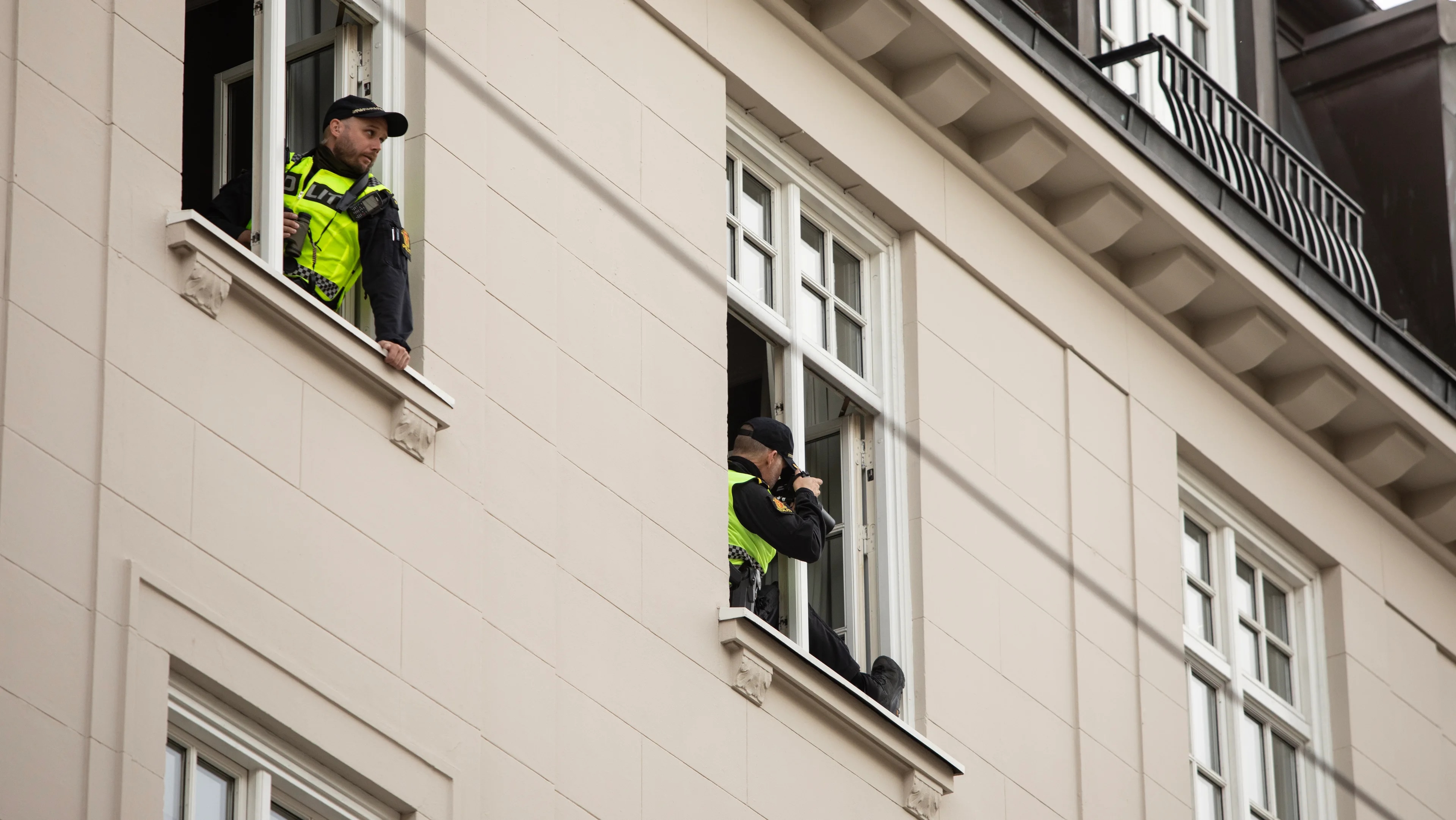To politibetjenter i uniform følger med fra hvert sitt vindu i en bygård. (Bildebeskrivelsen er laget av en KI-tjeneste)