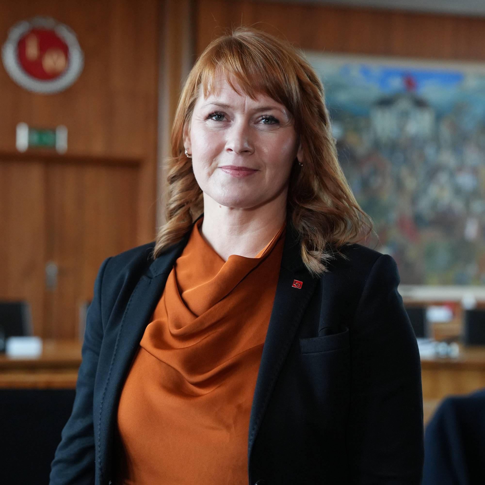 En kvinne i mørk dress og orange topp ser mot kamera. Bak henne er et møterom med panelkledde vegger og et maleri på veggen. En logo med rød sirkel og et symbol er synlig. (Bildebeskrivelsen er laget av en KI-tjeneste)