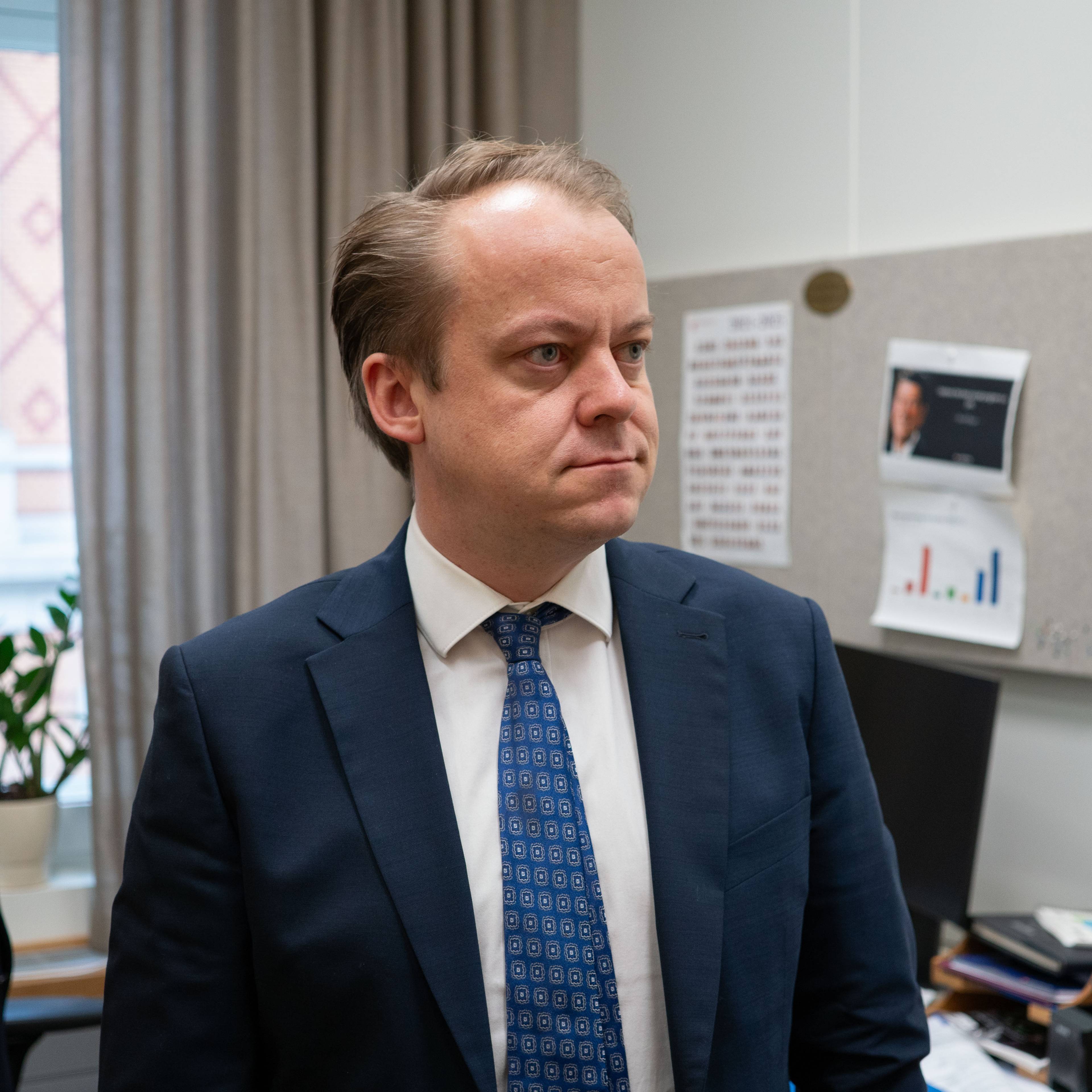 stortingsrepresentant for Fremskrittspartiet, Erlend Wiborg, står på sitt kontor. Han ser ut til siden og ser alvorlig ut. Han har på seg hvit skjorte, mørkeblå dress og blått slips med et mønster.