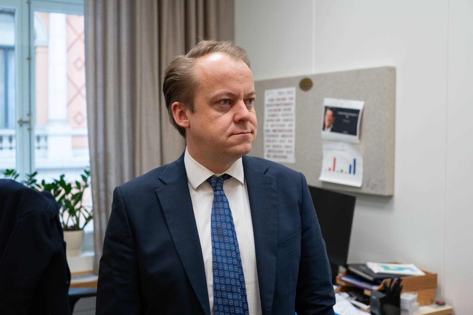 stortingsrepresentant for Fremskrittspartiet, Erlend Wiborg, står på sitt kontor. Han ser ut til siden og ser alvorlig ut. Han har på seg hvit skjorte, mørkeblå dress og blått slips med et mønster. 