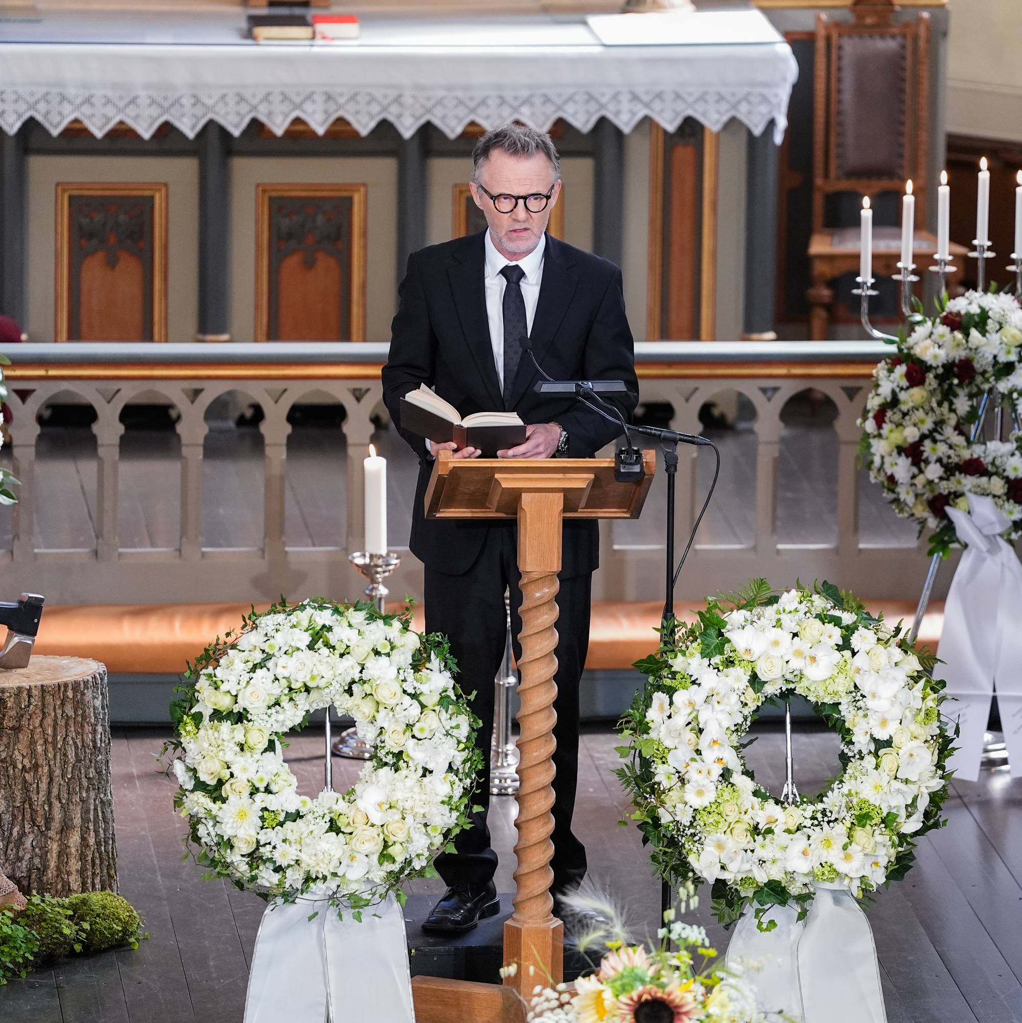 Mann i dress holder tale fra en talerstol i en kirke omgitt av blomsterkranser.