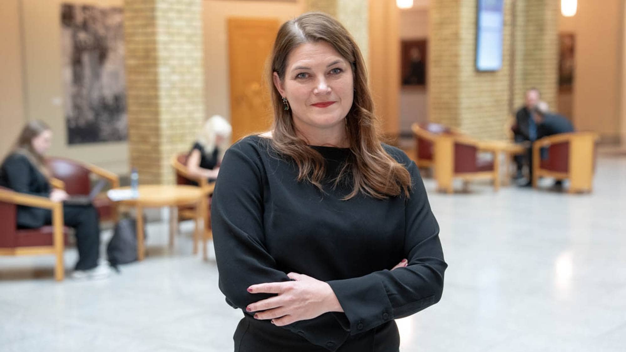 En kvinne med langt hår og svart klær står med armene krysset i en bygning. I bakgrunnen sitter flere personer som arbeider, mens noen har samtaler. Omgivelsene har en moderne arkitektur med mursteinsvegger og store vinduer. Det er et lyst og åpent rom med sitteplasser.